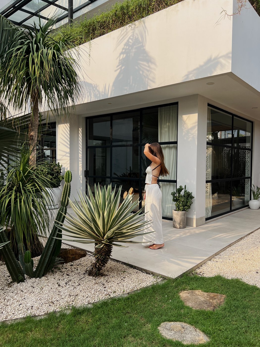 A woman stands outside a modern building surrounded by tropical plants, adjusting her hair in the sunlight.