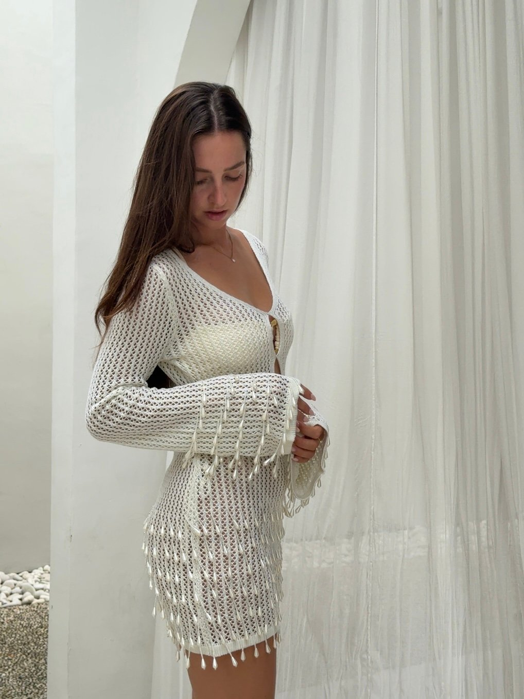 A woman in a white crochet dress with bell sleeves stands gracefully against a light backdrop, showcasing the intricate design of her outfit.