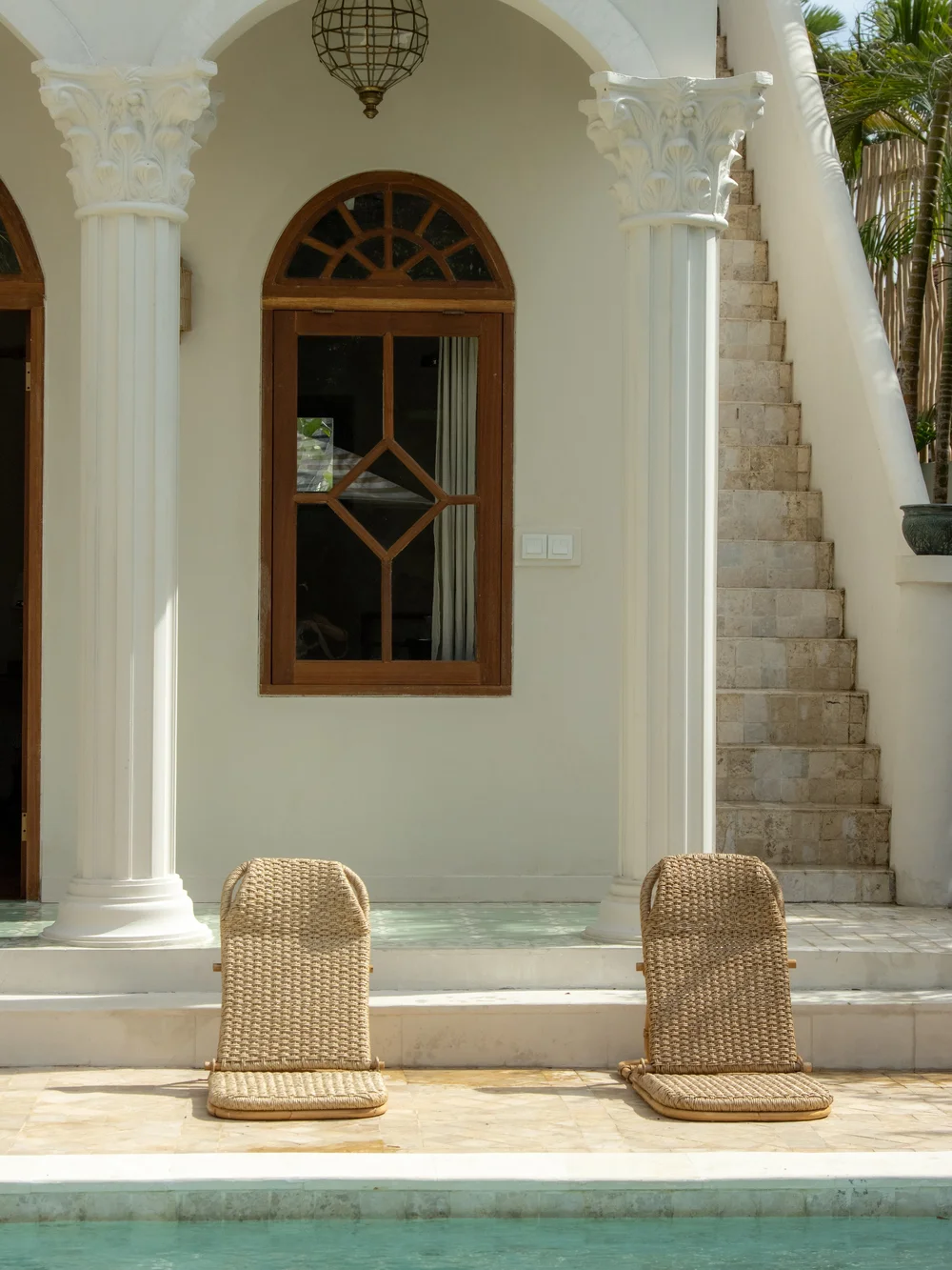 Two woven chairs positioned by a pool in front of a stylish villa with arched windows and a staircase.