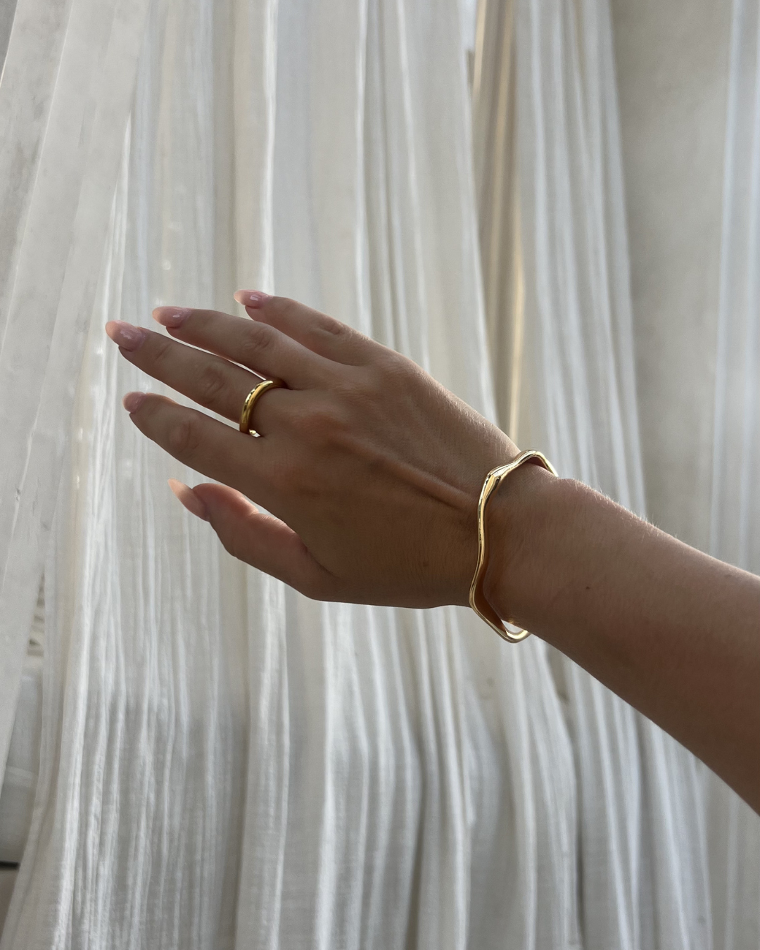 A close-up of a hand wearing a gold bracelet and ring, positioned against sheer white curtains.