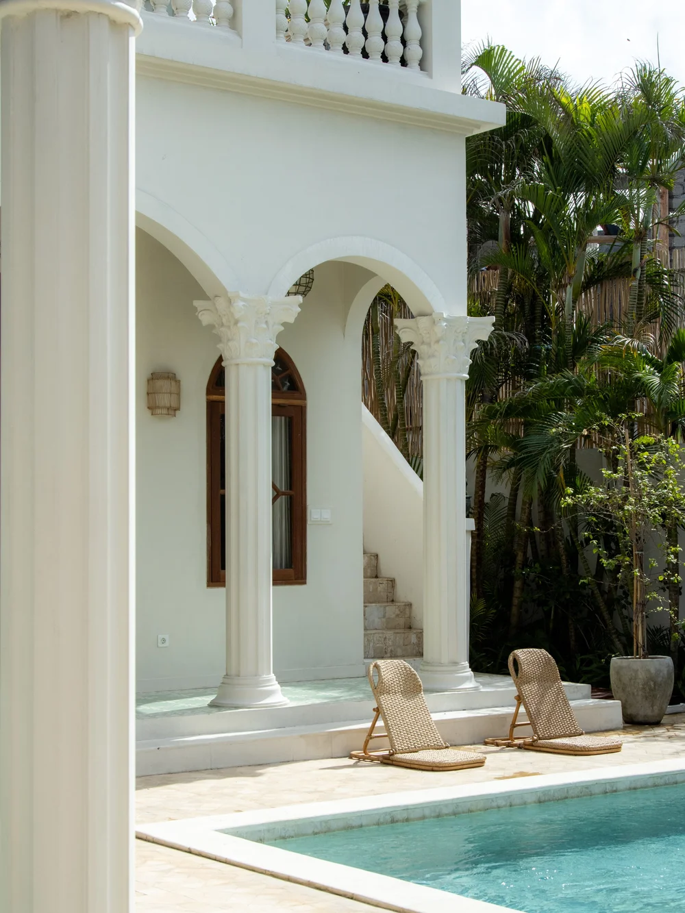 A serene outdoor space featuring a pool and two lounge chairs beside a white villa with elegant columns and lush greenery.