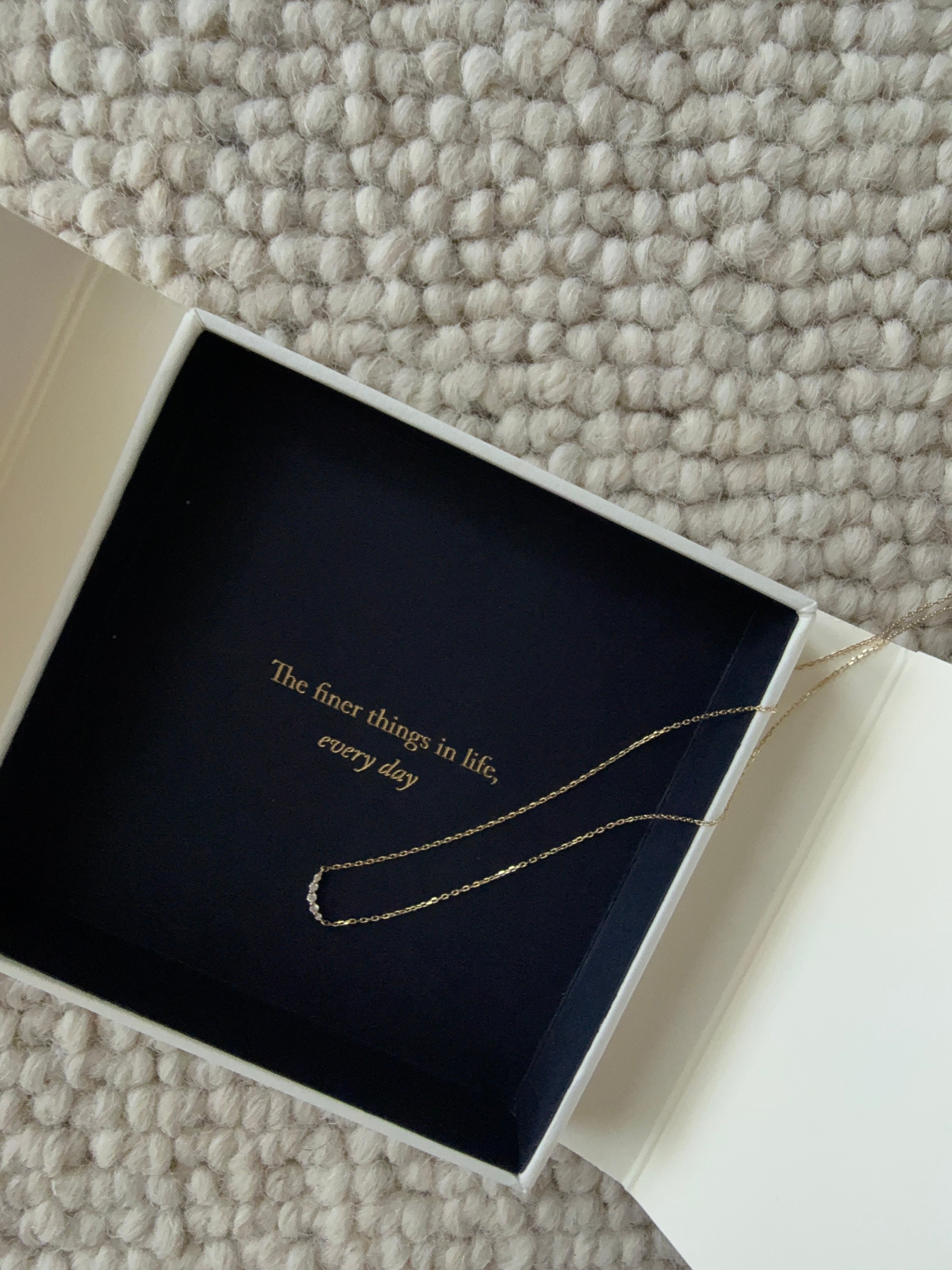 A delicate gold necklace displayed inside a jewelry box with a dark blue interior. The box features an elegant inscription that reads, 'The finer things in life, every day.'
