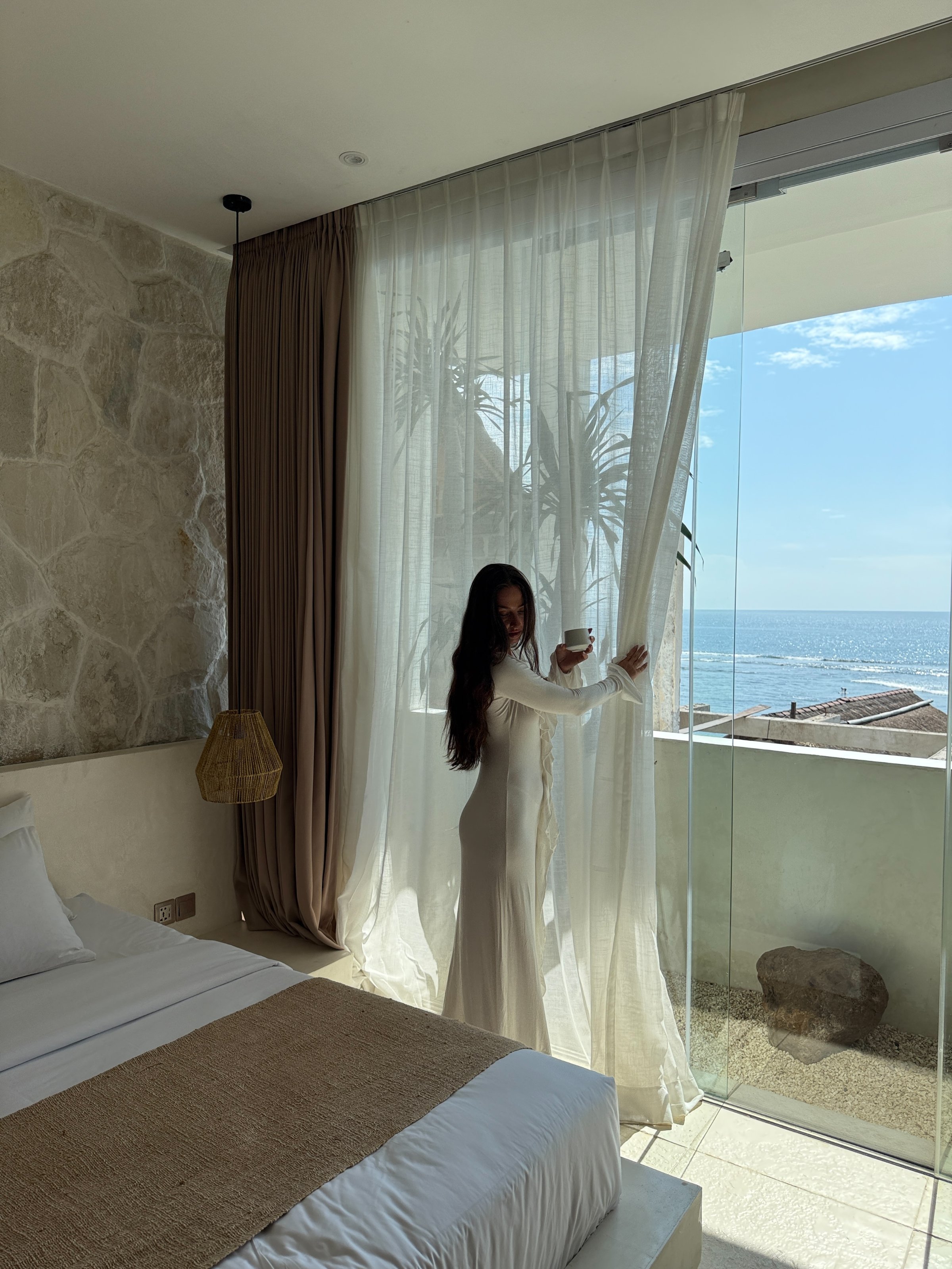 A person in a long white dress stands by a window, pulling back sheer curtains to reveal a view of the ocean.