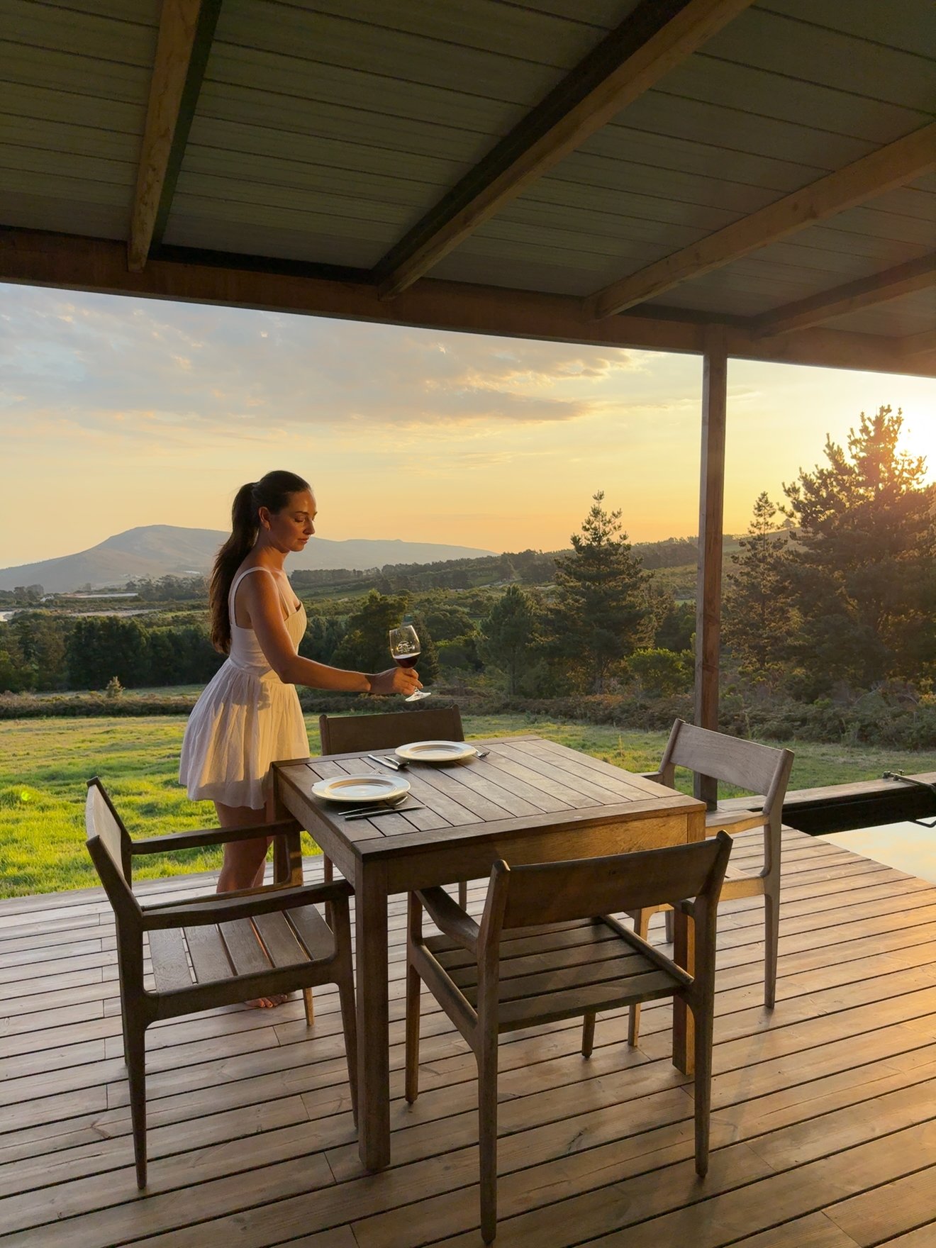 A woman in a white dress stands at a wooden table on a porch, holding a glass of wine while overlooking a scenic sunset landscape.