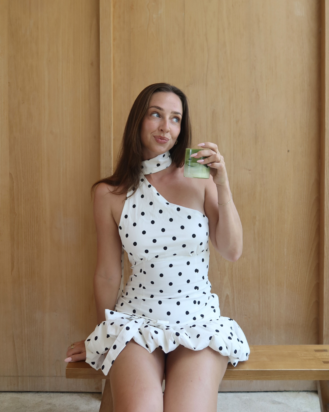 A woman in a polka dot dress sits on a wooden bench, holding a green drink and looking playfully at the camera.
