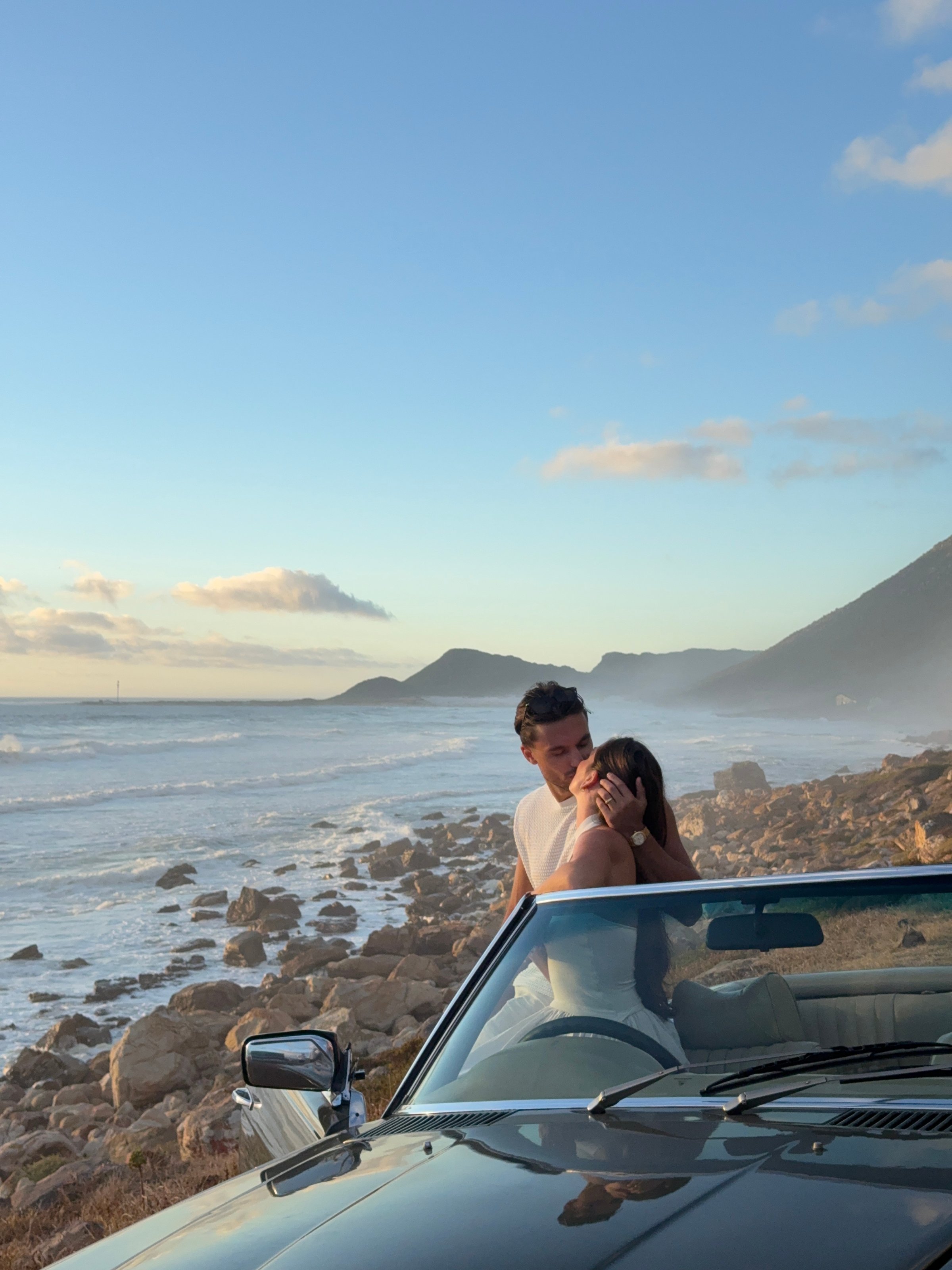 A couple embraces in a convertible car by the beach, with mountains and a cloudy sky in the background.