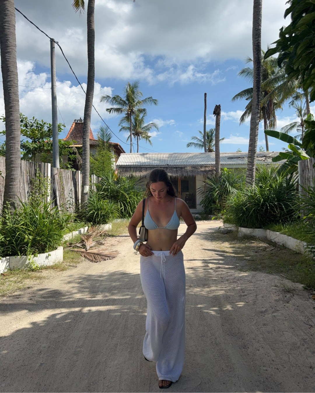 A woman walks down a sandy path lined with palm trees and greenery, wearing a bikini top and white pants, under a partly cloudy sky.