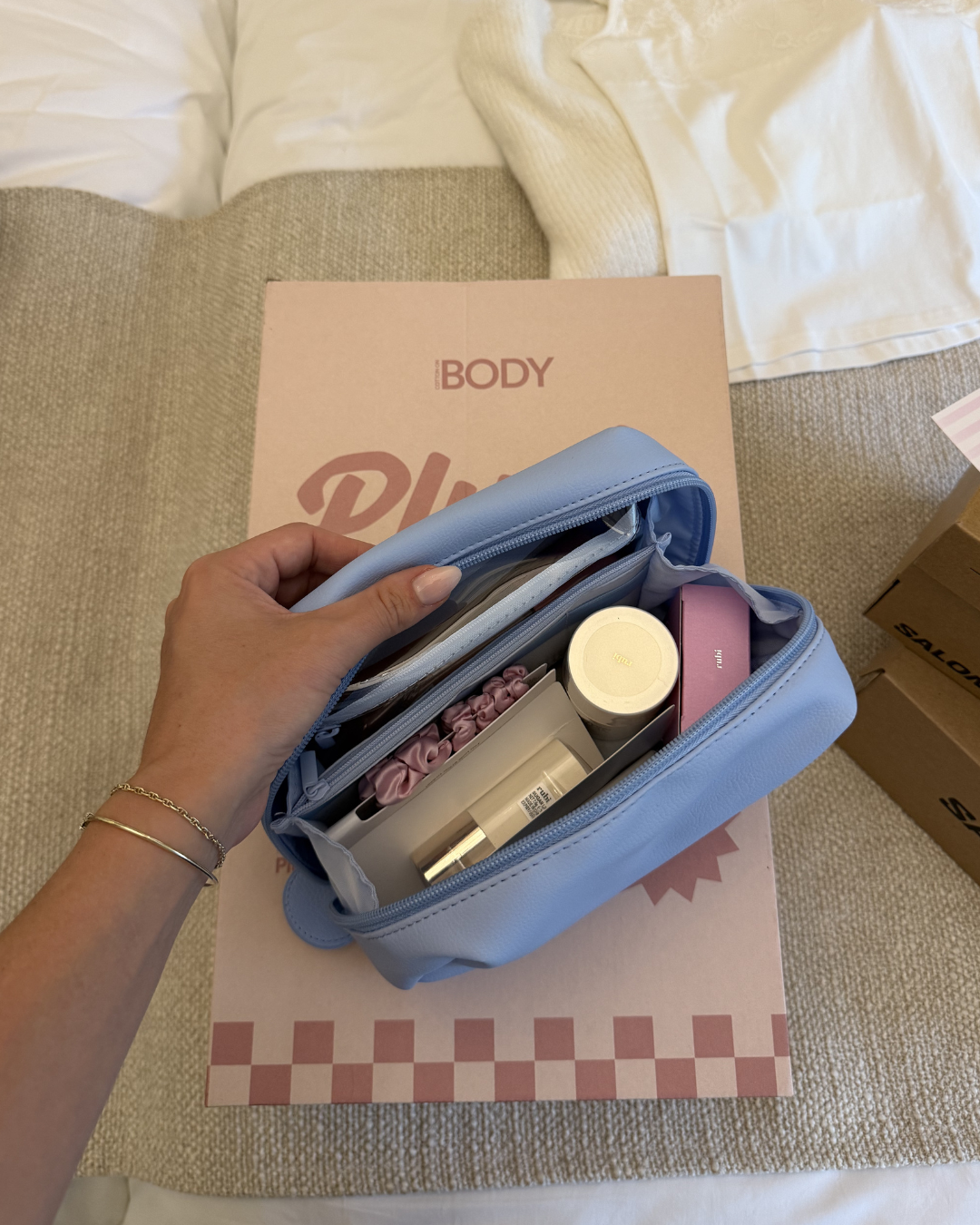 A hand holding a light blue cosmetic bag filled with various beauty products, placed on a pink box with the text 'iBODY'.