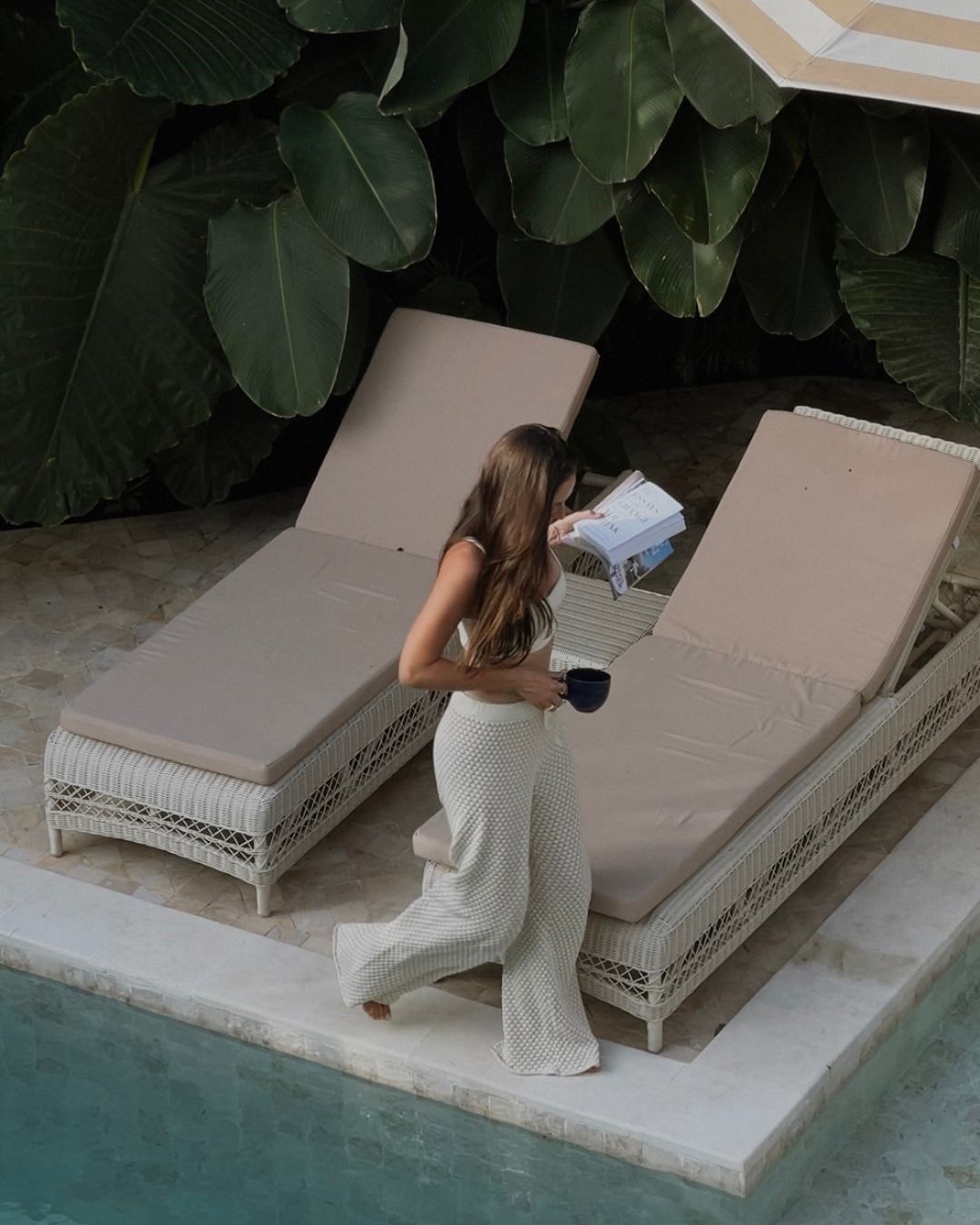 A woman walks by a poolside while holding a book and a cup, surrounded by lush green foliage.