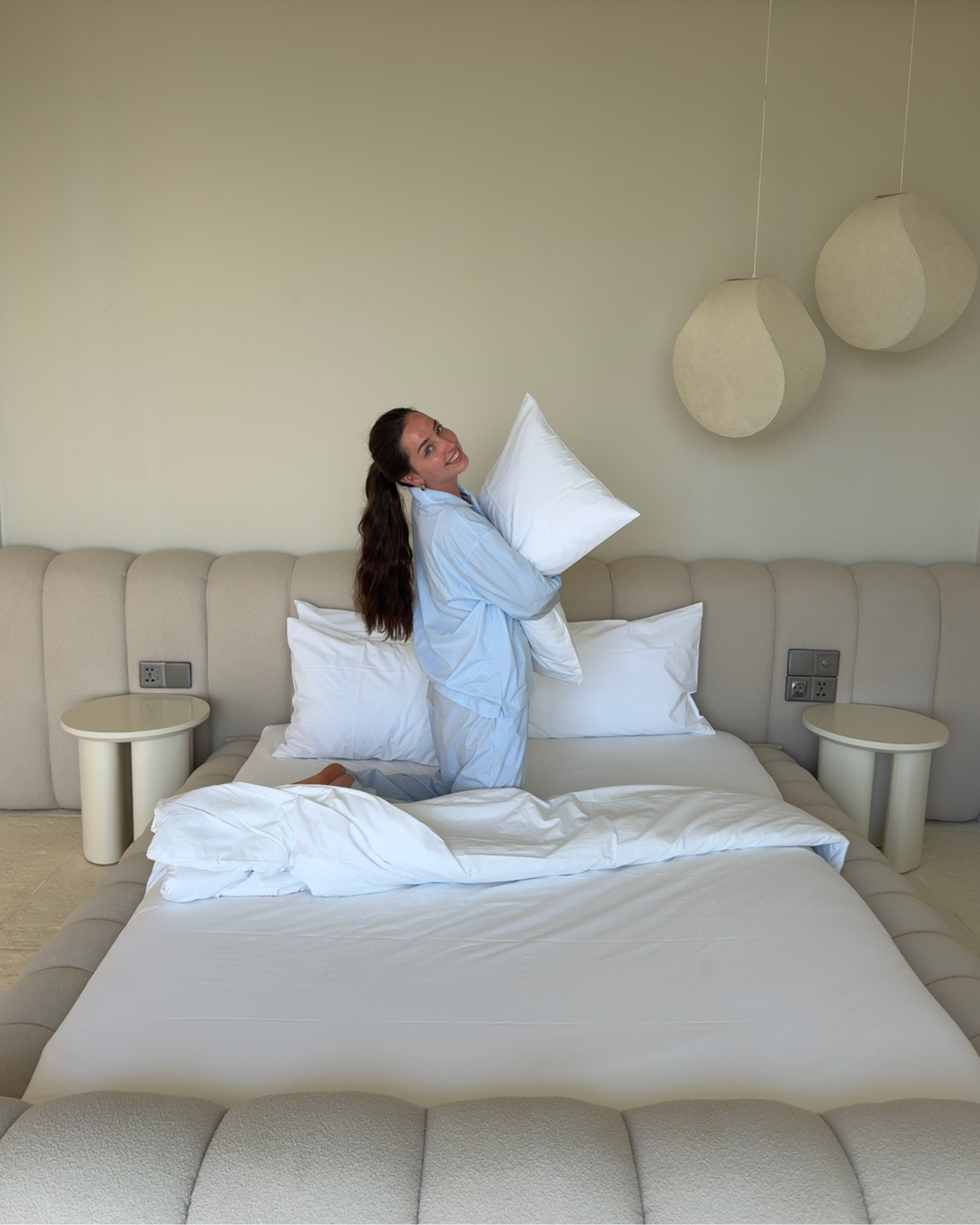 A person in light blue pajamas playfully holds a pillow while sitting on a neatly made bed in a modern room.