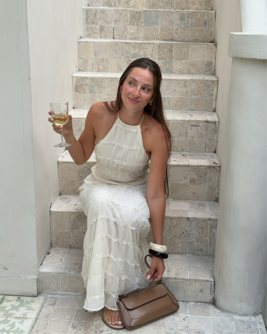 A woman in a white dress sitting on a staircase, holding a glass of drink and smiling. She has long hair and is accessorized with a bracelet and a handbag.