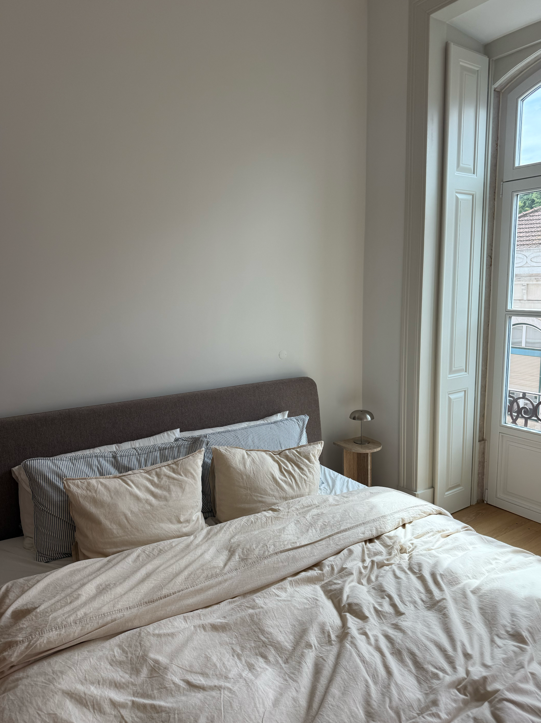 A cozy bedroom featuring a neatly made bed with soft pillows and a light-colored duvet, illuminated by natural light from a large window.