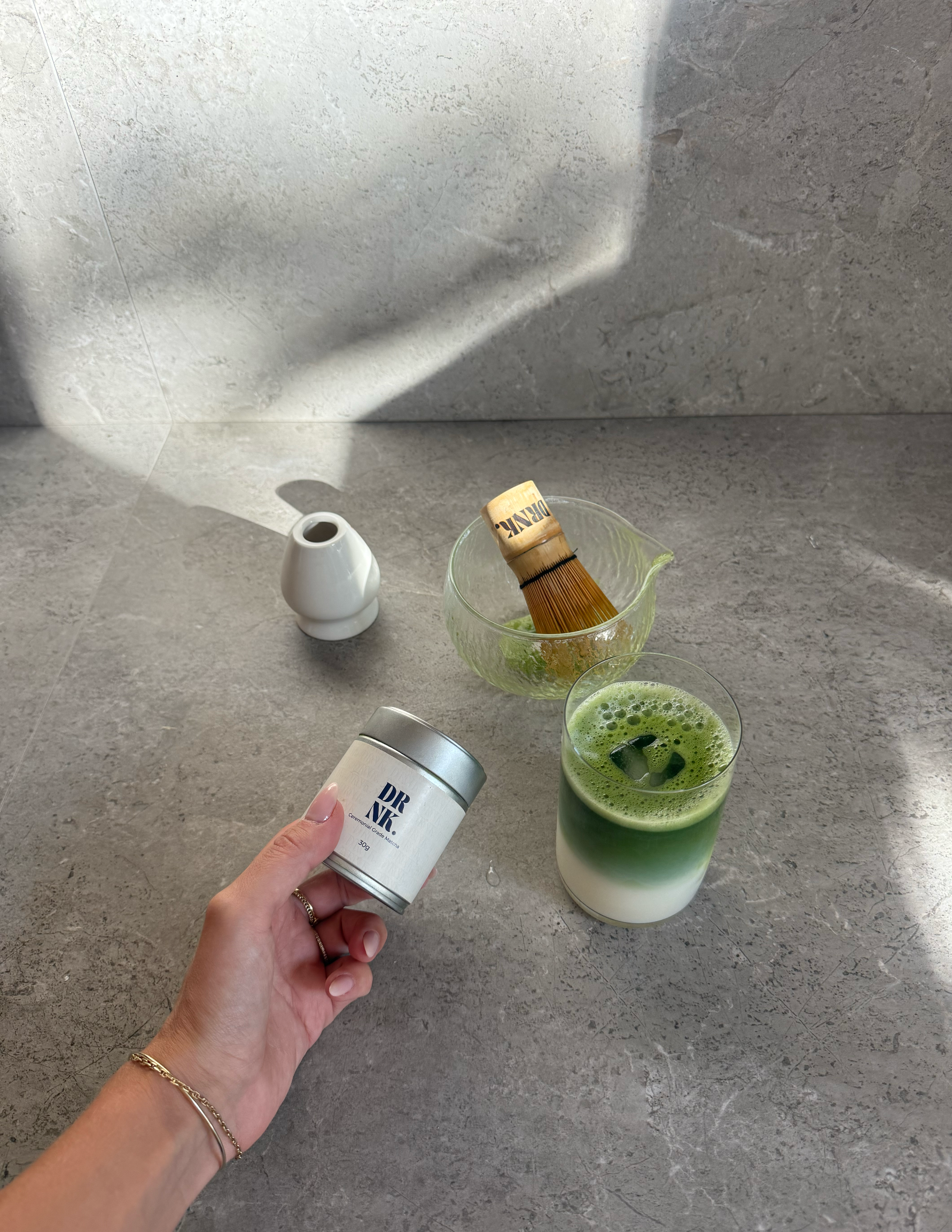 A hand holding a jar labeled 'DR AIR' next to a glass of layered green beverage, a bamboo whisk, and a small white vase on a textured surface.