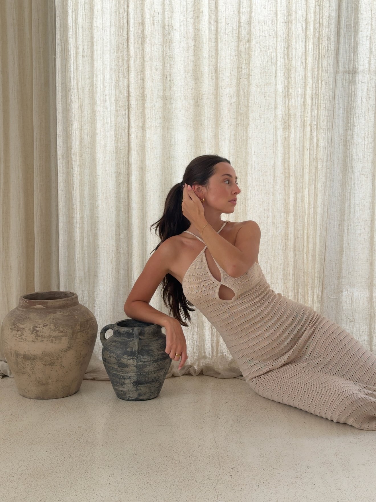 A woman in a knitted dress poses elegantly beside two decorative pots against a backdrop of sheer curtains.