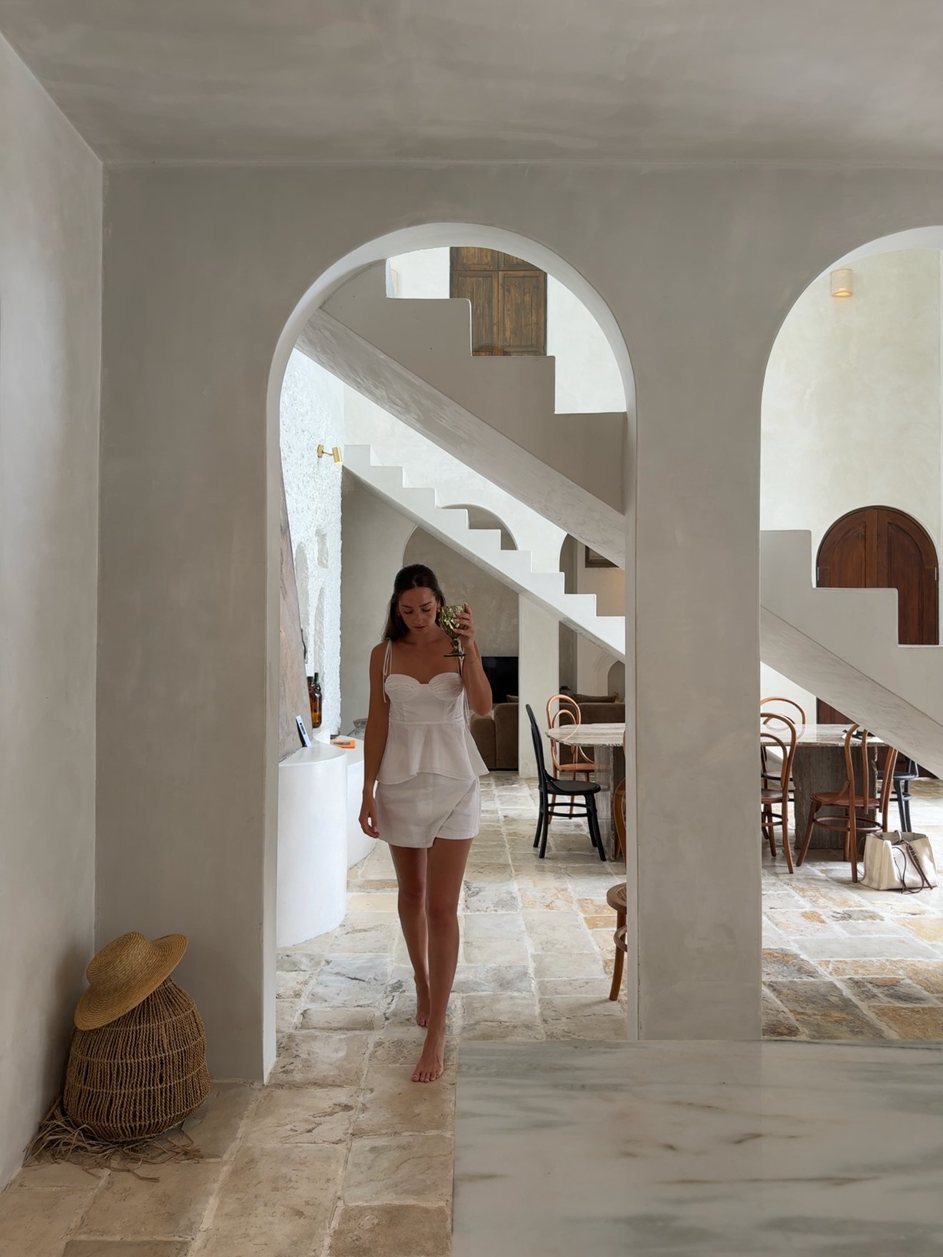 A woman stands barefoot in a modern, minimalist interior with arched doorways and a staircase in the background. The space features natural materials and a warm color palette.