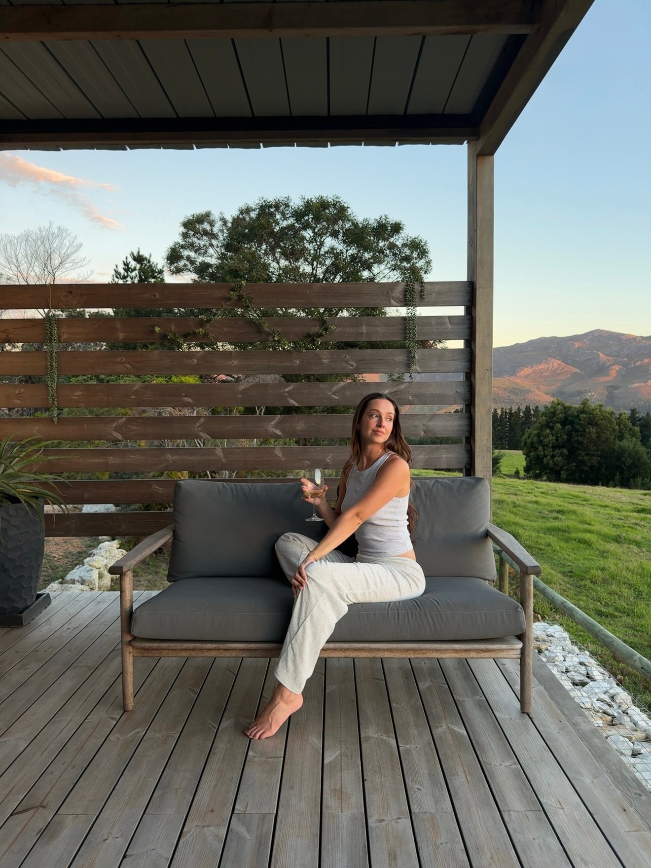 A woman sitting on a wooden bench on a porch, holding a drink, with a scenic view of mountains and greenery in the background.