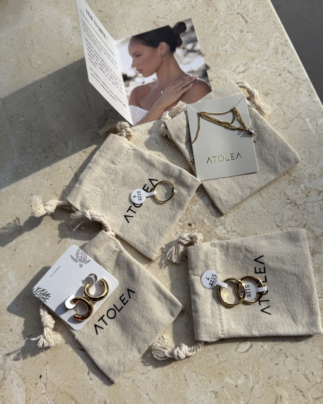 A collection of jewelry pieces displayed on a marble surface, featuring small fabric pouches and a promotional card with a model wearing the jewelry.