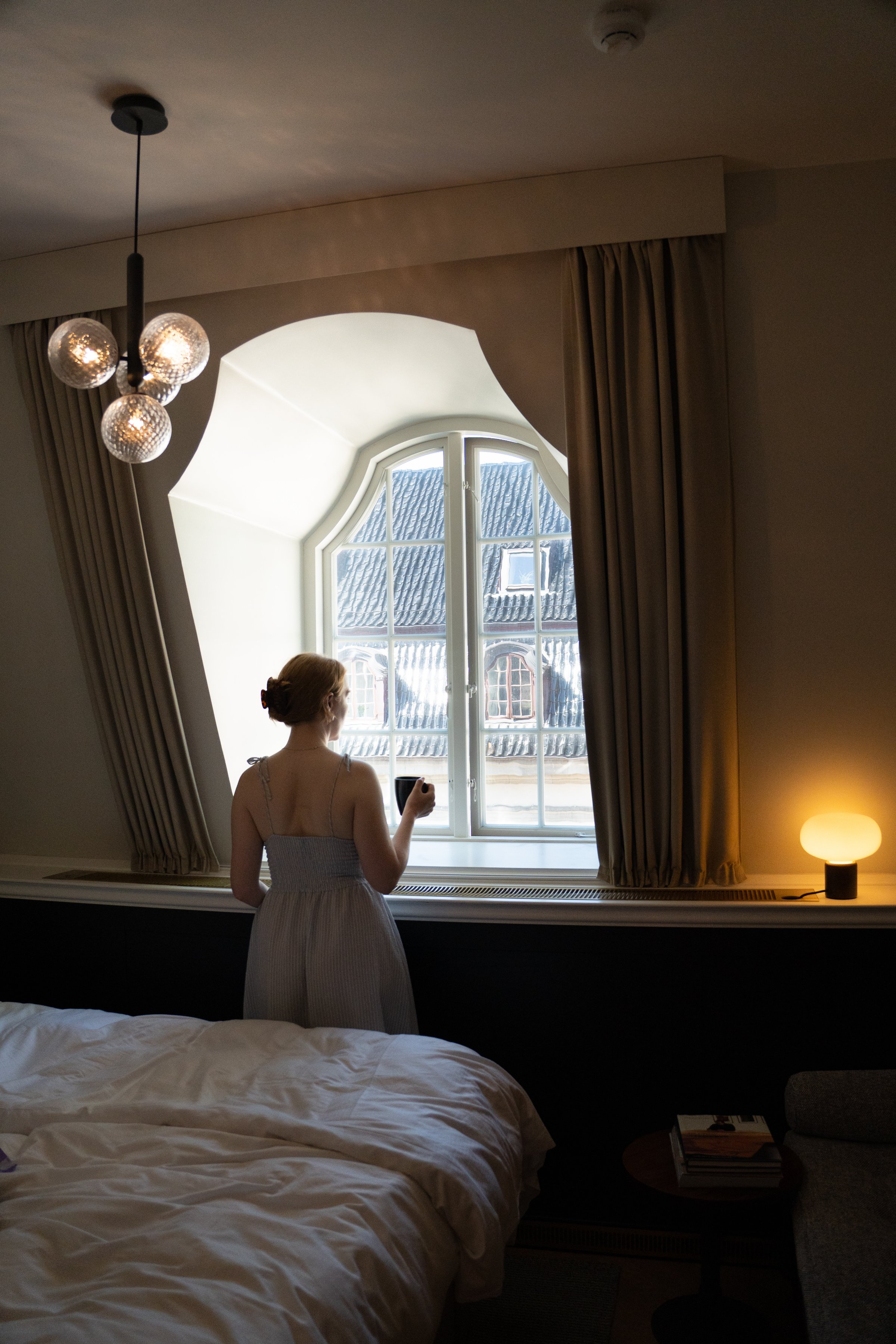 A woman in a light dress stands by a large window, holding a cup and looking outside. The room features soft lighting and a cozy atmosphere.