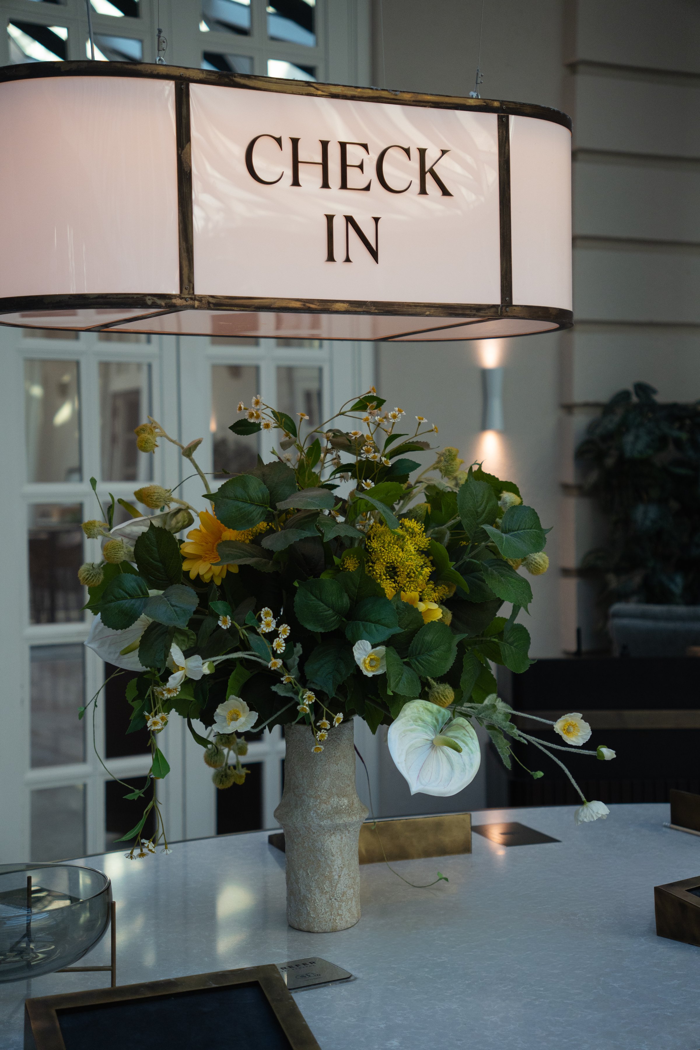 A decorative arrangement of flowers in a vase placed on a table, with a sign above reading 'CHECK IN'.
