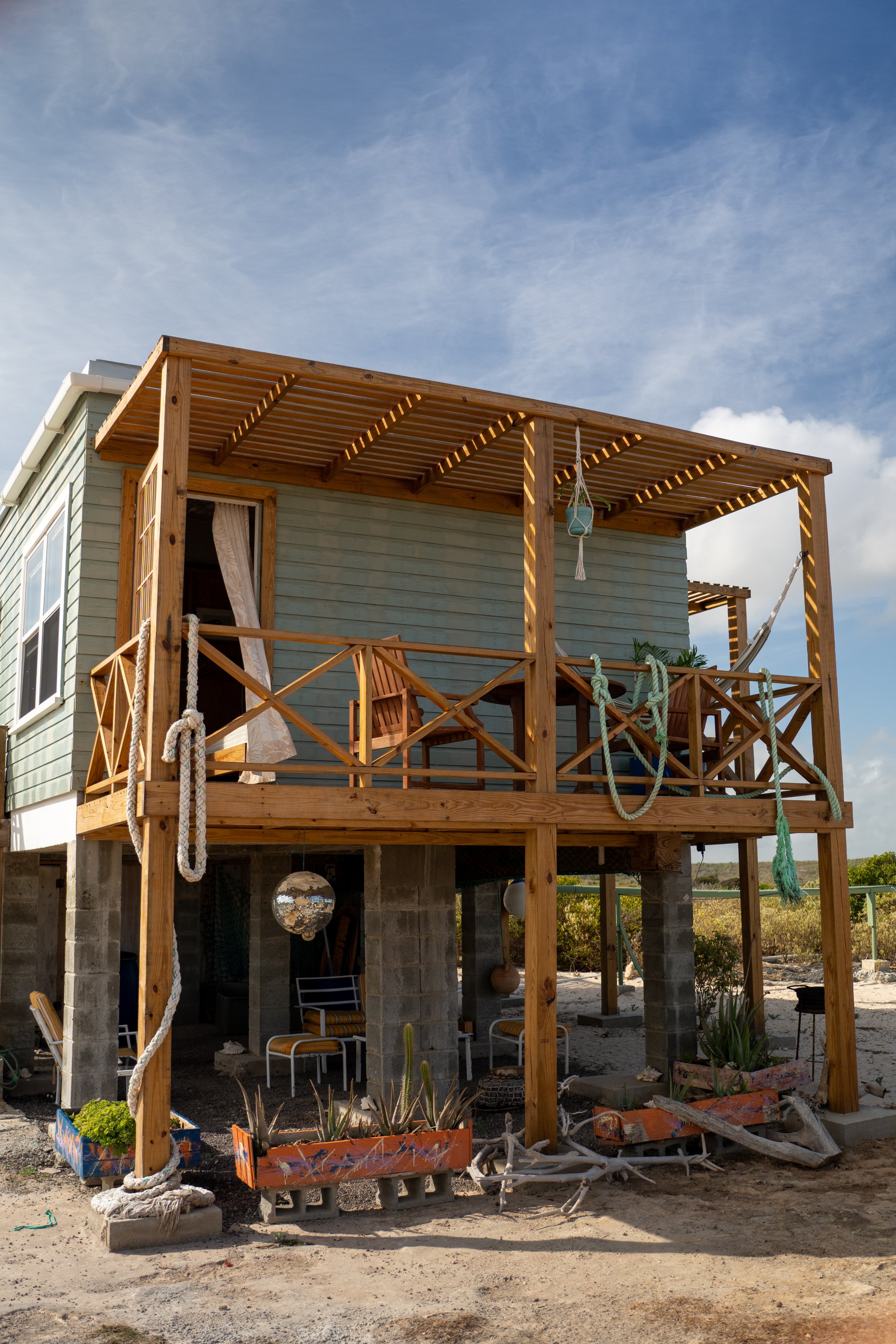 A wooden house on stilts with a spacious deck, featuring chairs and ropes, set against a blue sky.