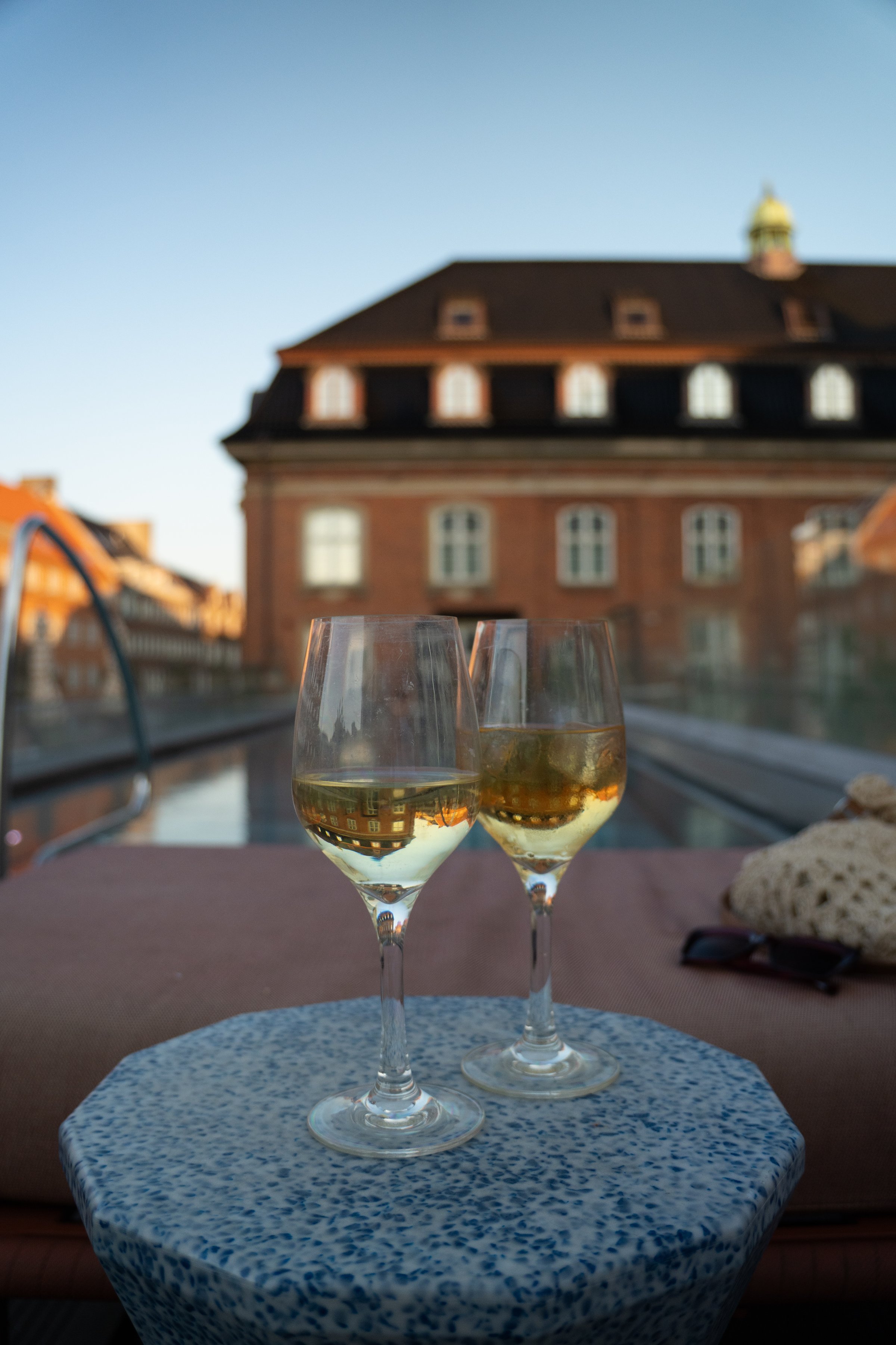 Two glasses of sparkling wine on a patterned table, with a scenic view of a building and a pool in the background.