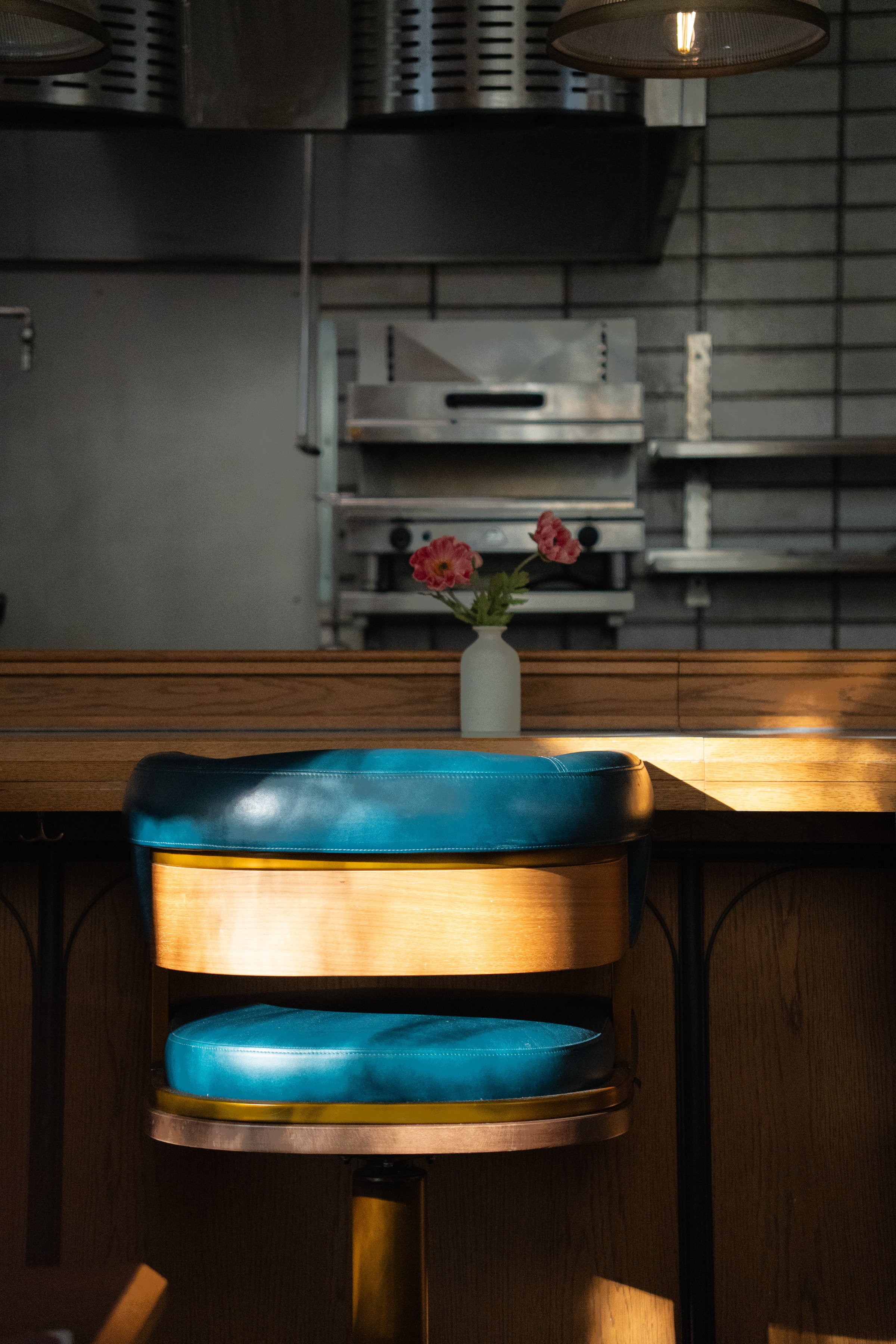 A close-up view of a blue bar stool with wooden accents, positioned at a counter with a small vase of pink flowers in the background.