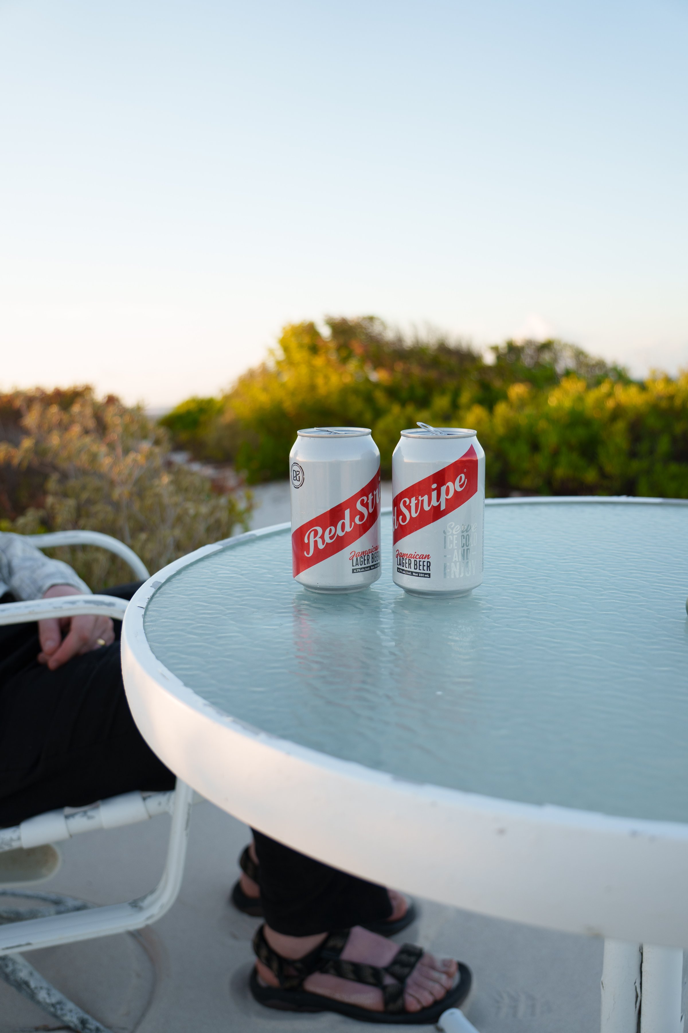 Two cans of Red Stripe beer sit on a glass table with a person’s legs visible in the background, surrounded by greenery and a clear sky.
