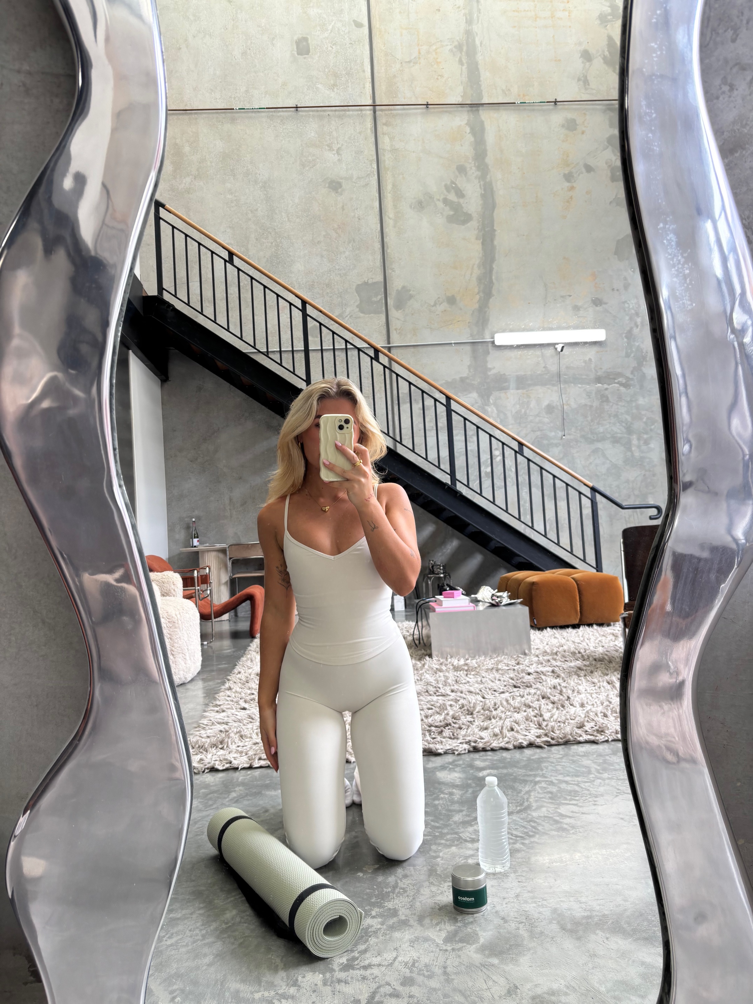 A person in a white outfit kneels on a yoga mat in a modern interior space with a large mirror and a staircase in the background.