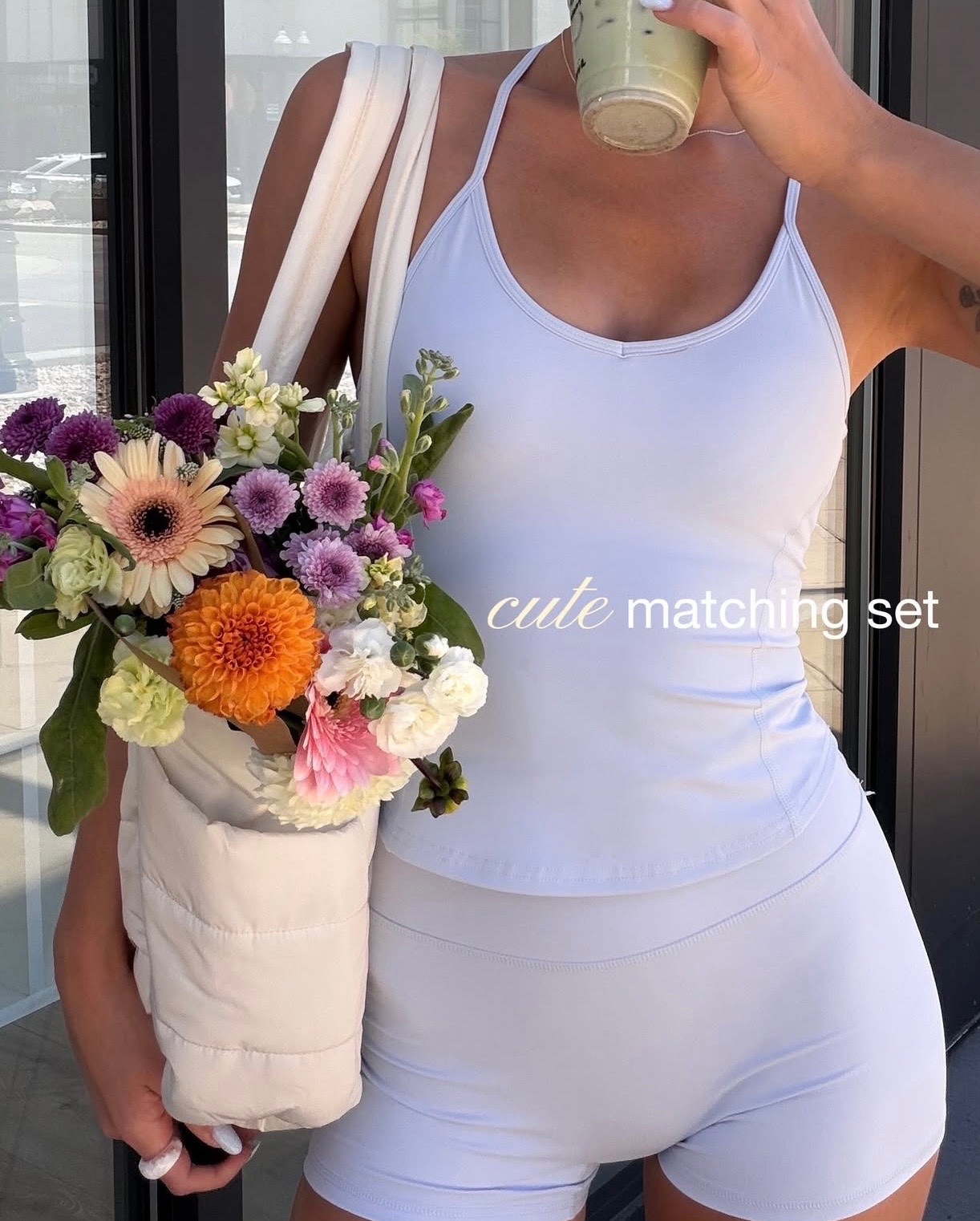 A person wearing a light purple matching athletic set holds a smoothie in one hand and a bag filled with colorful flowers in the other, standing outside a building.