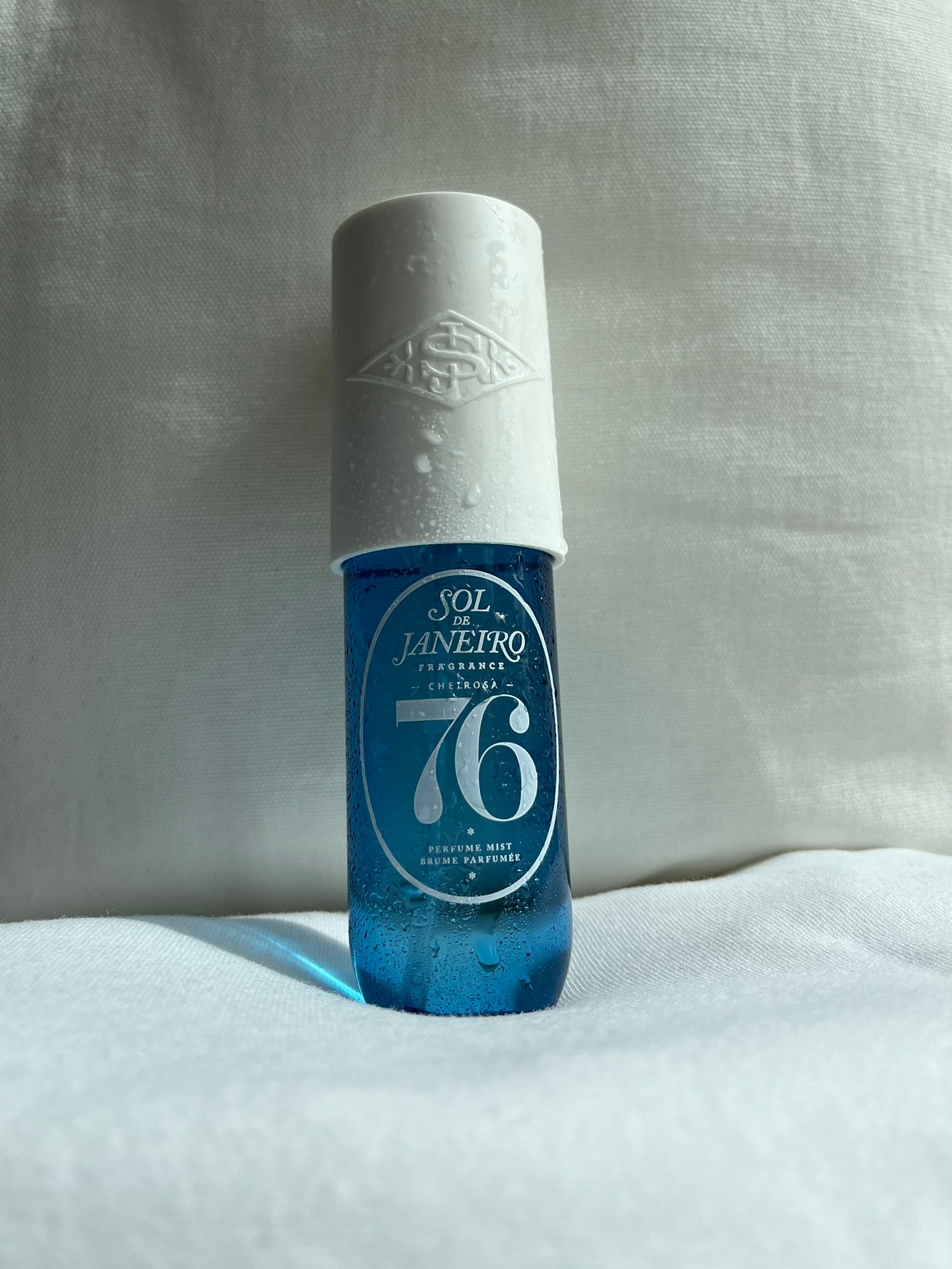 A blue bottle of Sol Janeiro fragrance with a white cap, placed on a light-colored fabric background.