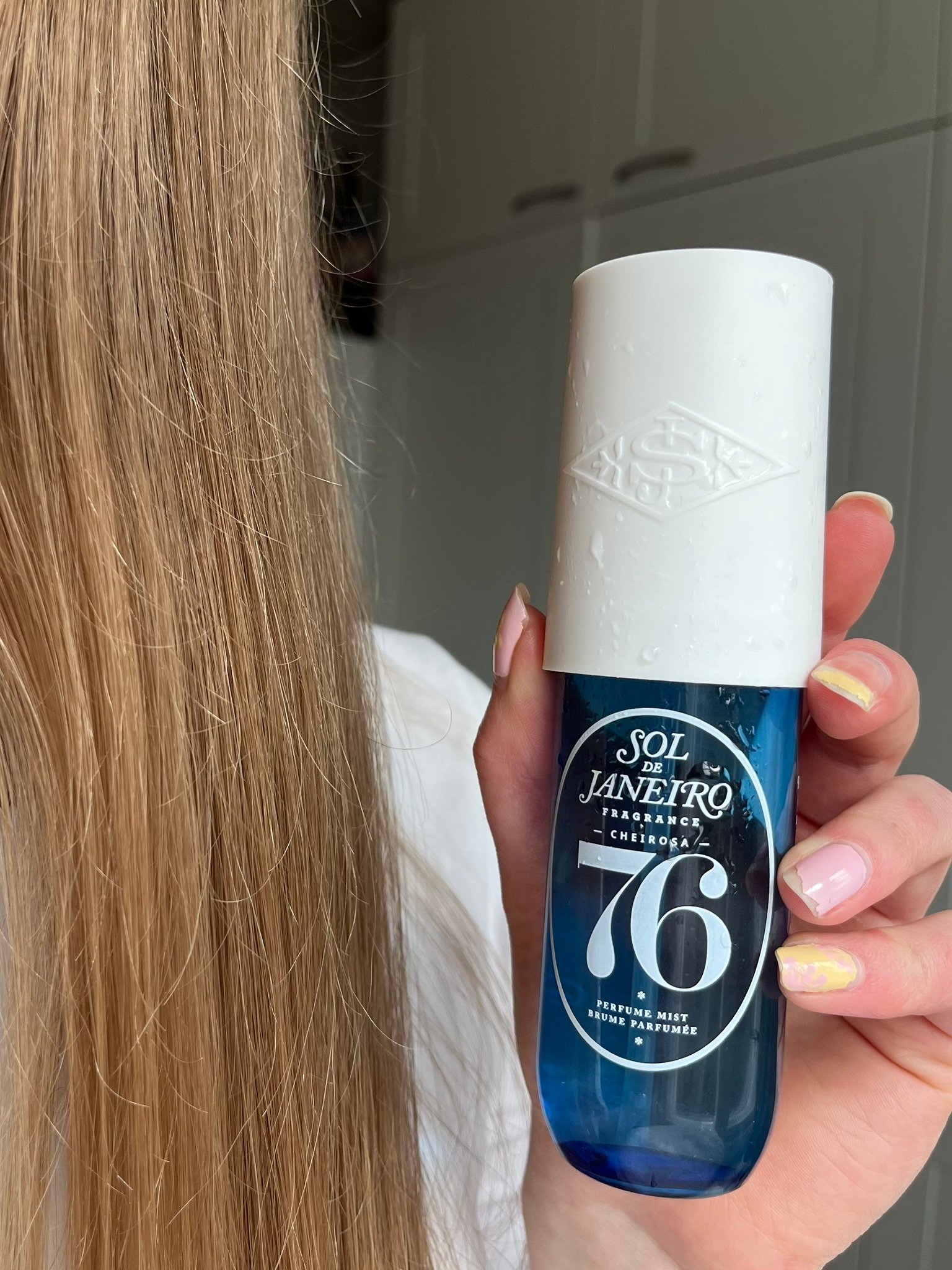 A person holding a blue bottle of Sol de Janeiro hair fragrance with a white cap, showcasing the product's label.