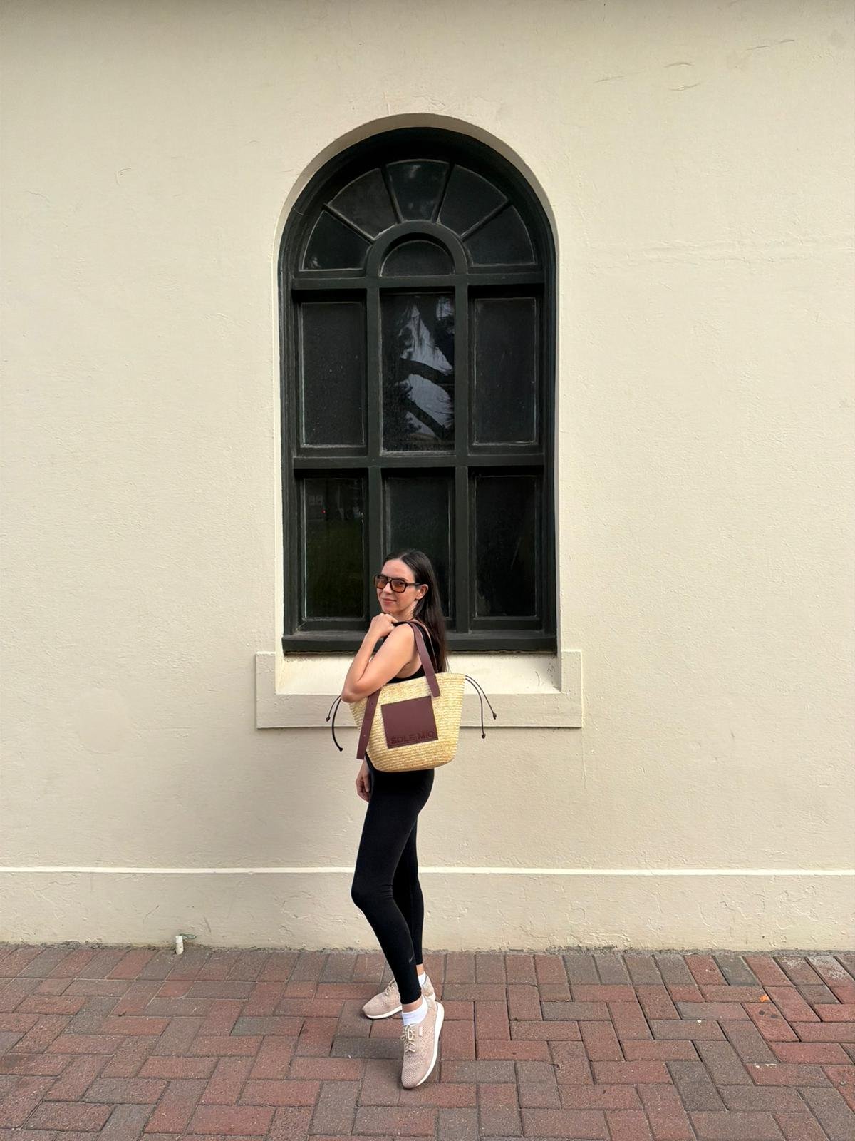 A woman stands confidently beside a large arched window, wearing black leggings and a stylish outfit, holding a woven bag.