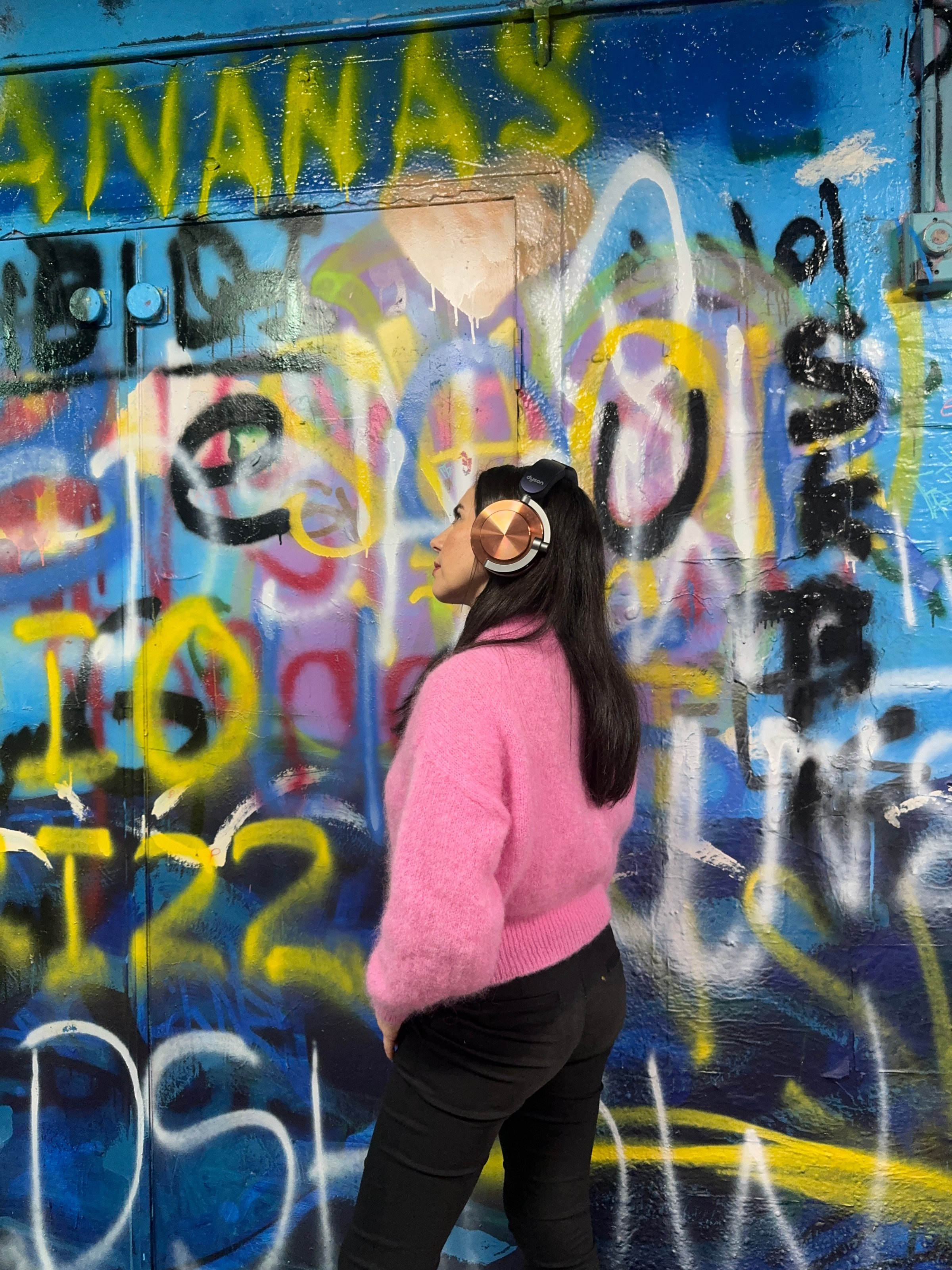 A person wearing headphones stands in front of a colorful graffiti wall, showcasing a vibrant urban backdrop.