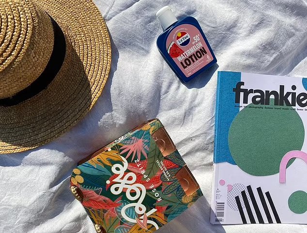 A flat lay arrangement featuring a straw hat, a bottle of sunscreen lotion, a colorful pouch, and a magazine on a white fabric surface.