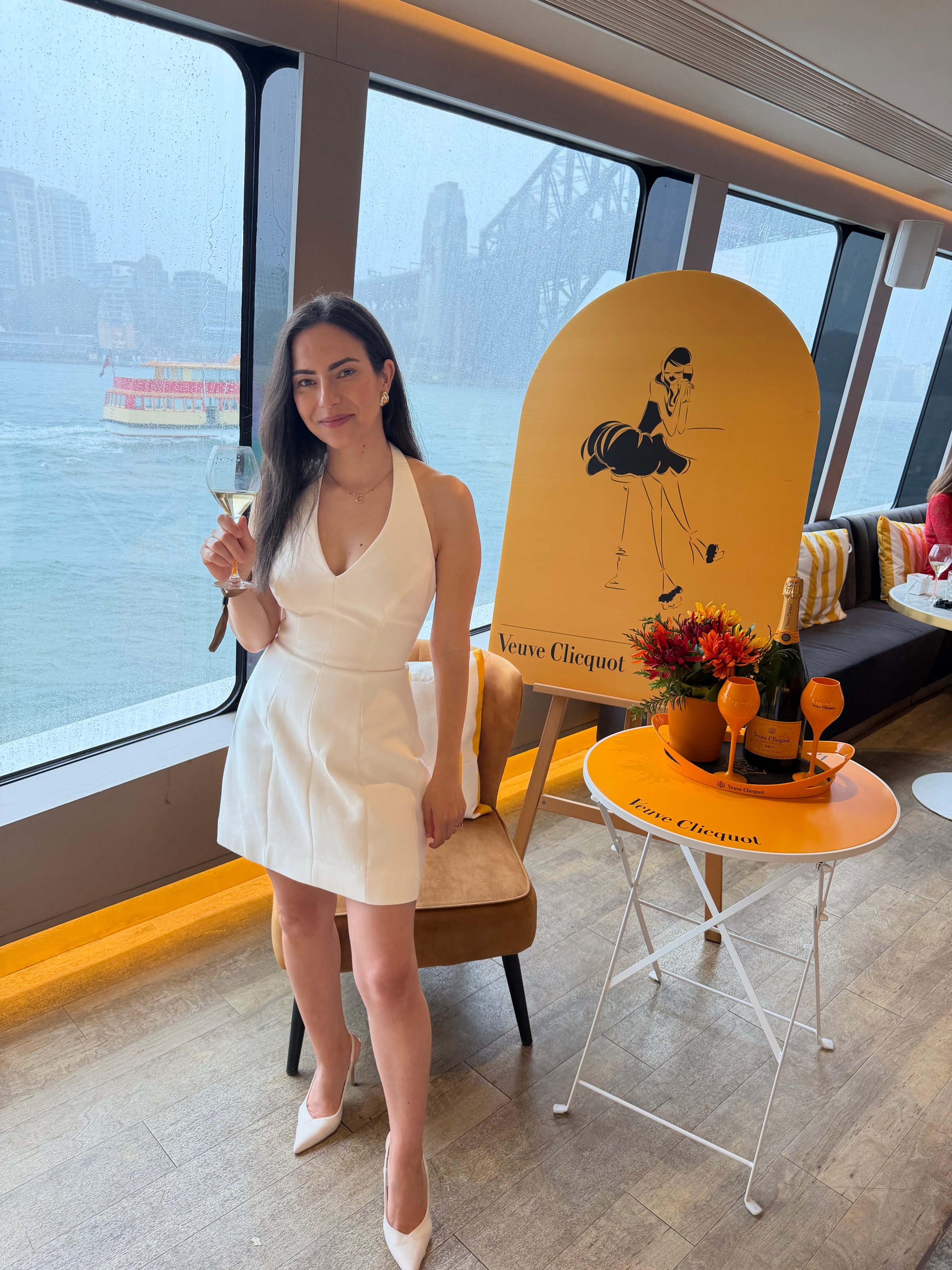A woman in a white dress holding a champagne glass, standing next to a table with flowers and drinks, with a view of a rainy cityscape in the background.