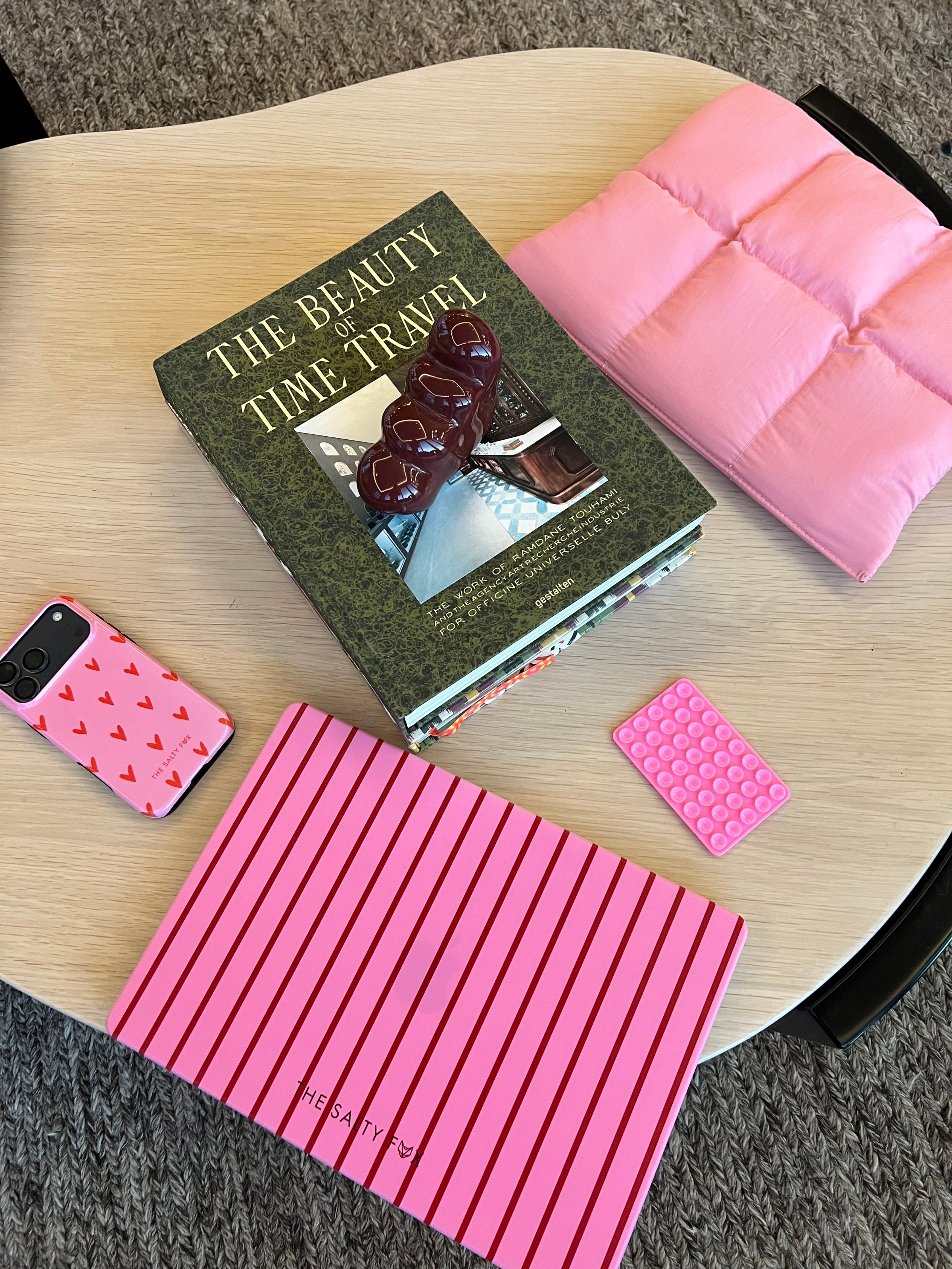 A flat lay of a coffee table featuring a book titled 'The Beauty of Time Travel', a pink cushion, a pink striped notebook, and a phone case with a heart pattern.