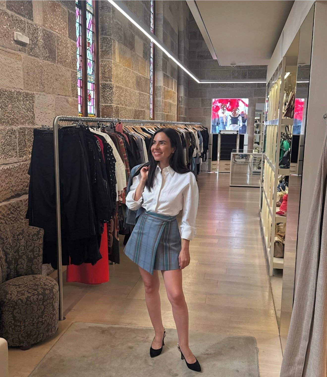A woman stands smiling in a stylish boutique, wearing a white shirt and a plaid skirt, with clothing racks in the background.
