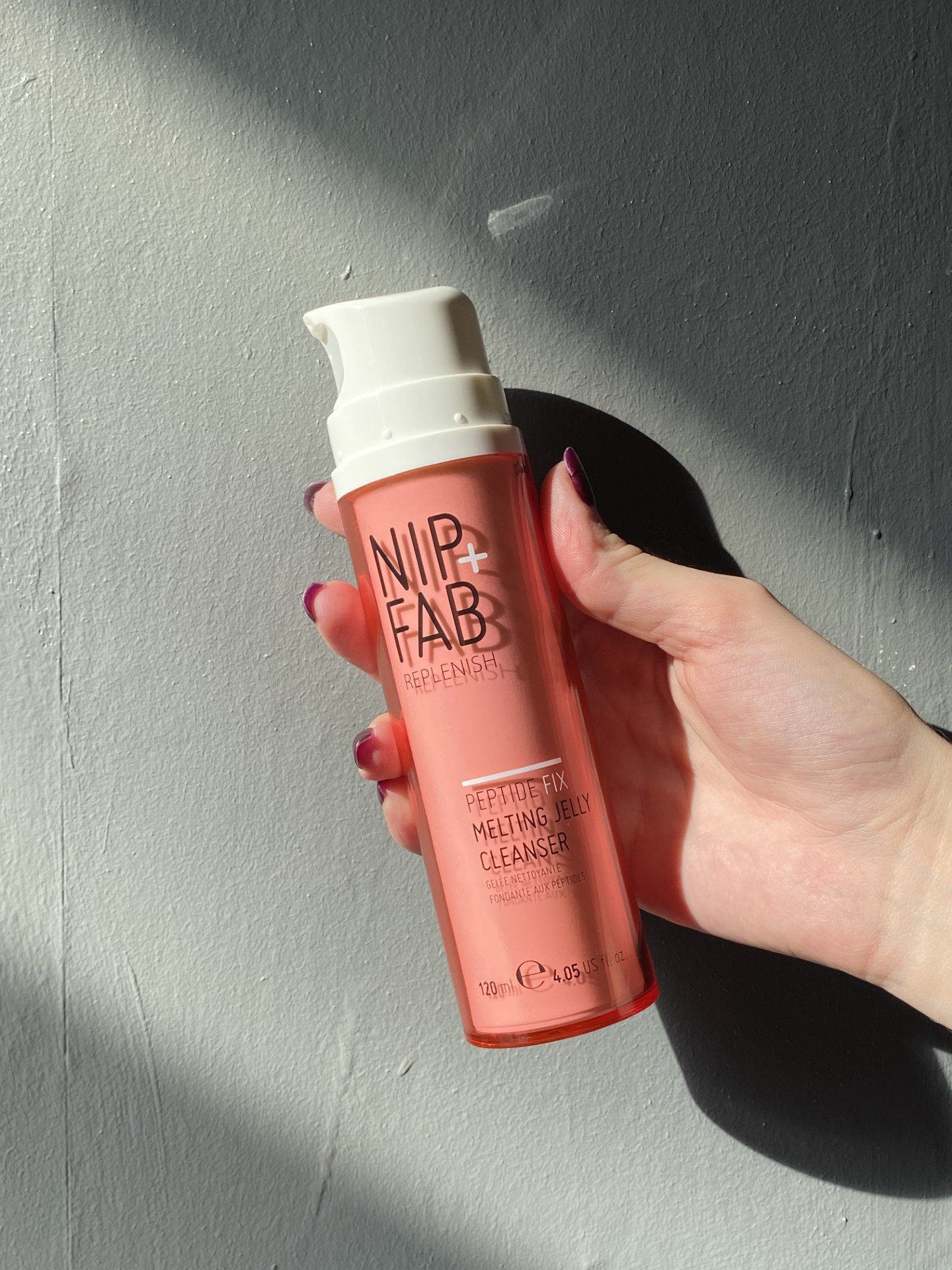 A hand holding a bottle of NIP+FAB cleansing gel against a textured gray wall.