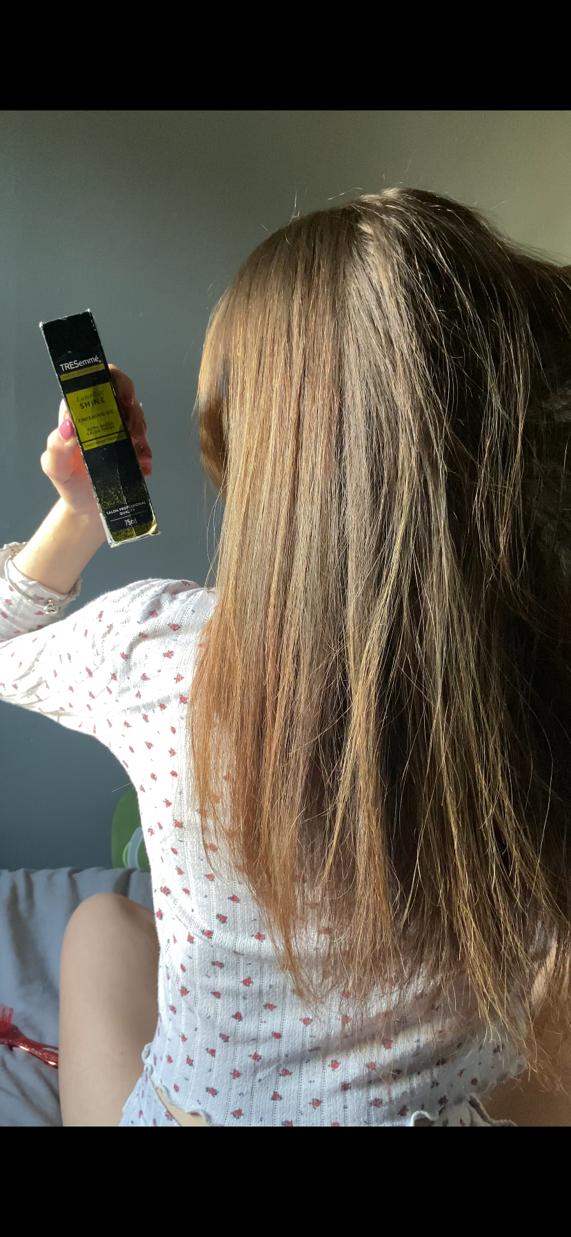 A person with long, straight hair is holding a hair product while sitting on a bed. The focus is on the hair and the product being applied.