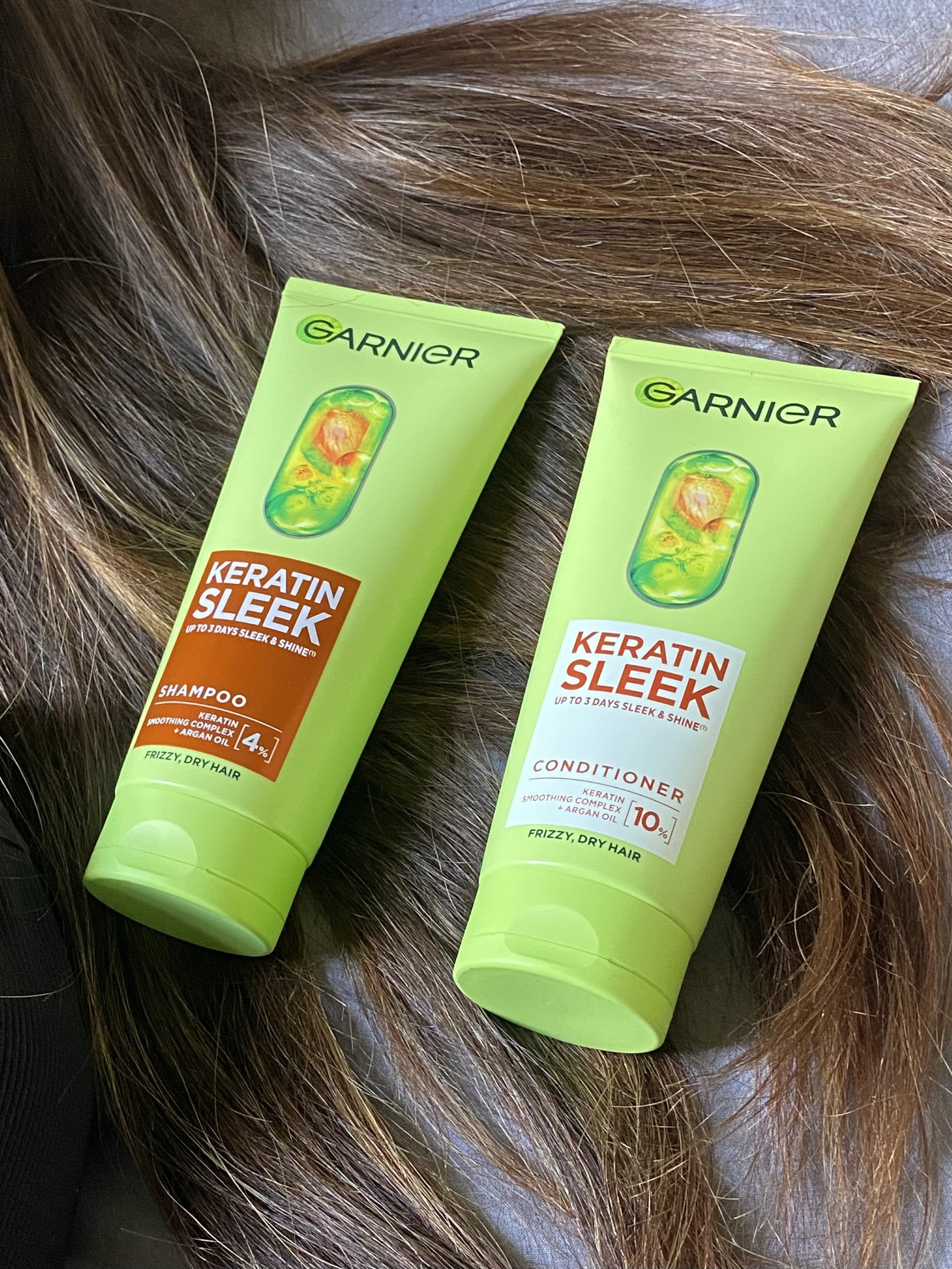 Two tubes of Garnier Keratin Sleek shampoo and conditioner placed on a textured surface with hair in the background.