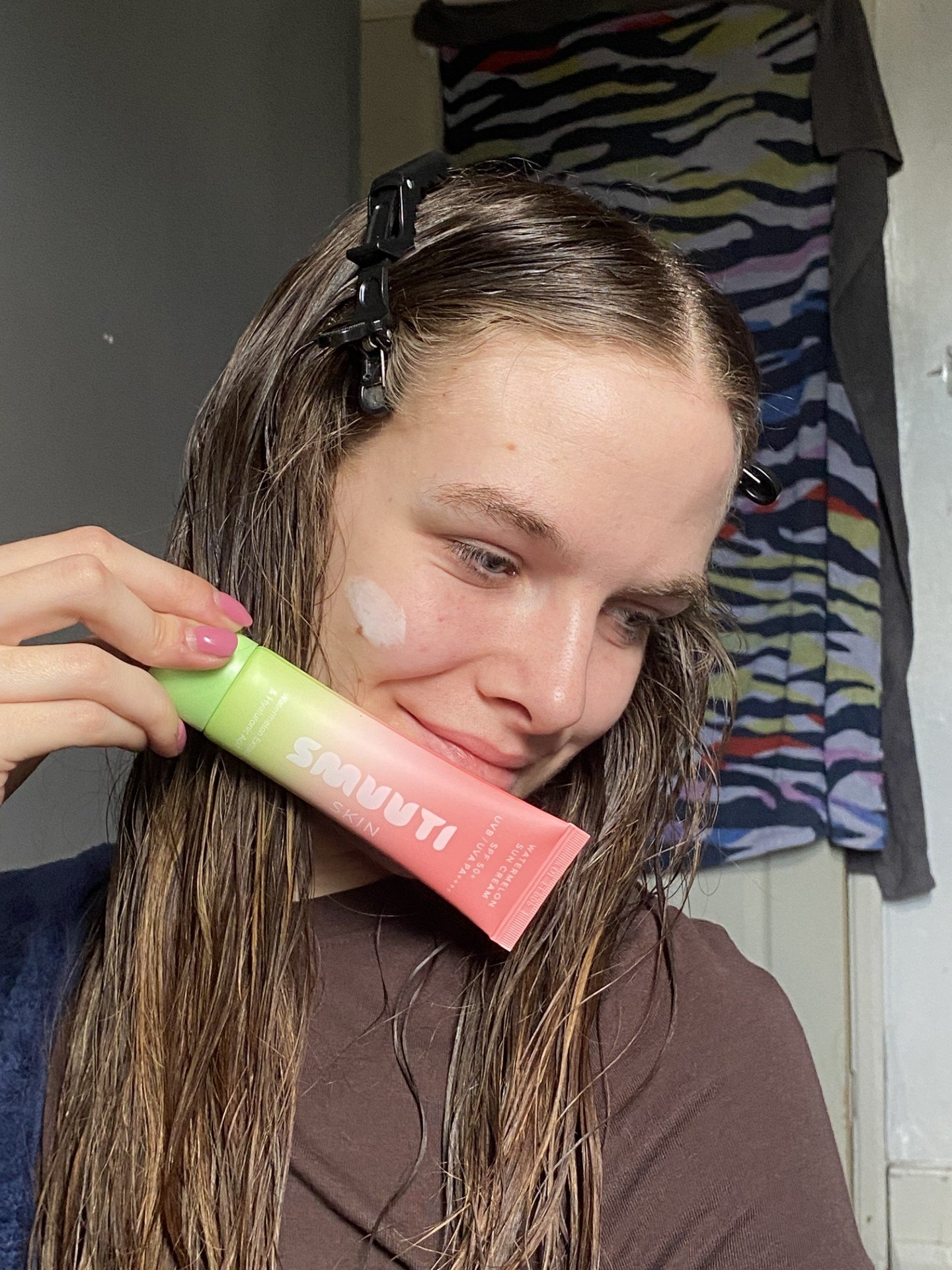 A person with wet hair holds a skincare product, smiling while applying it to their face.