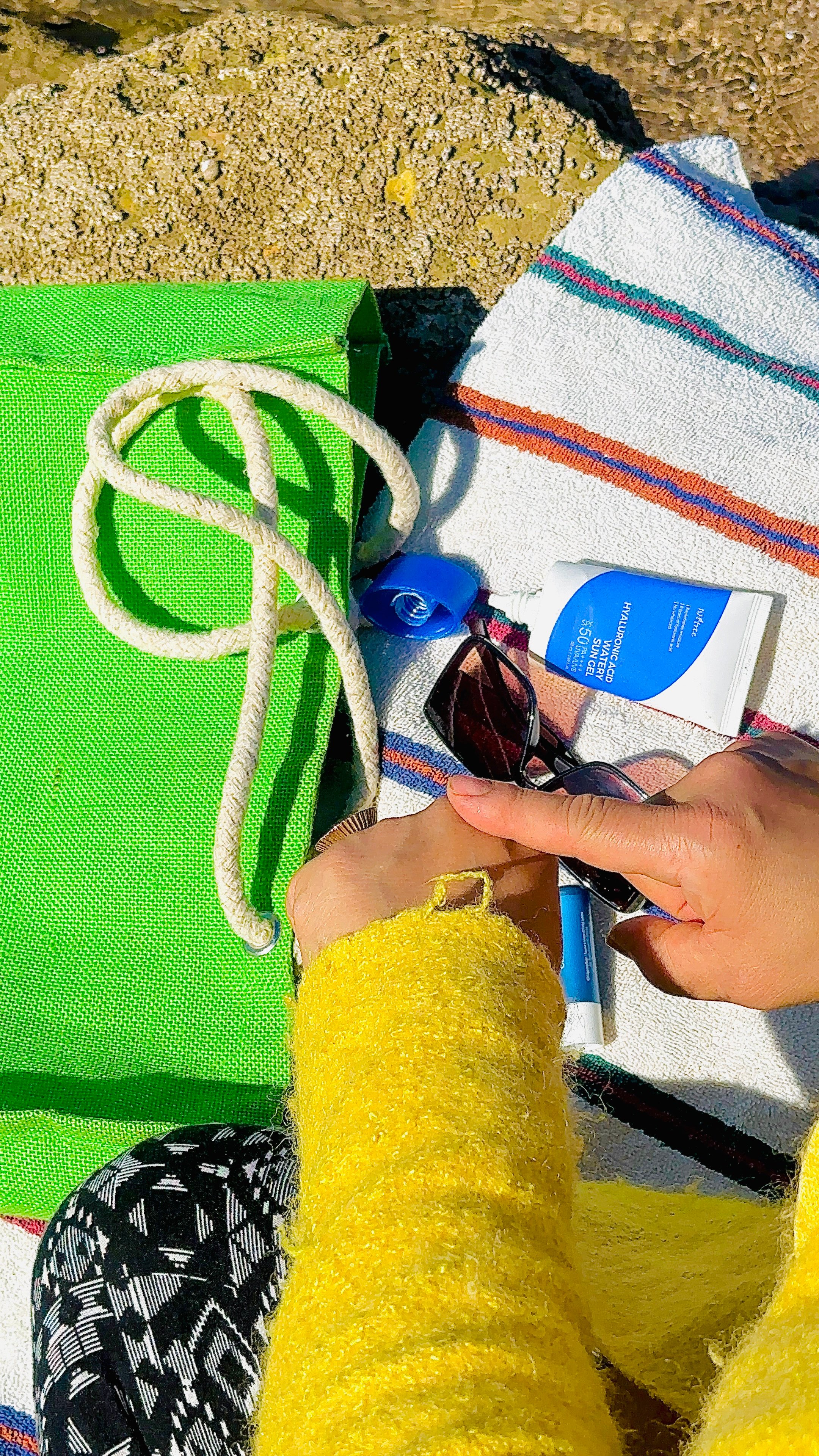 A person wearing a bright yellow sweater is sitting on a striped towel, with a green bag and sunglasses nearby. Sunscreen and a pen are also visible.
