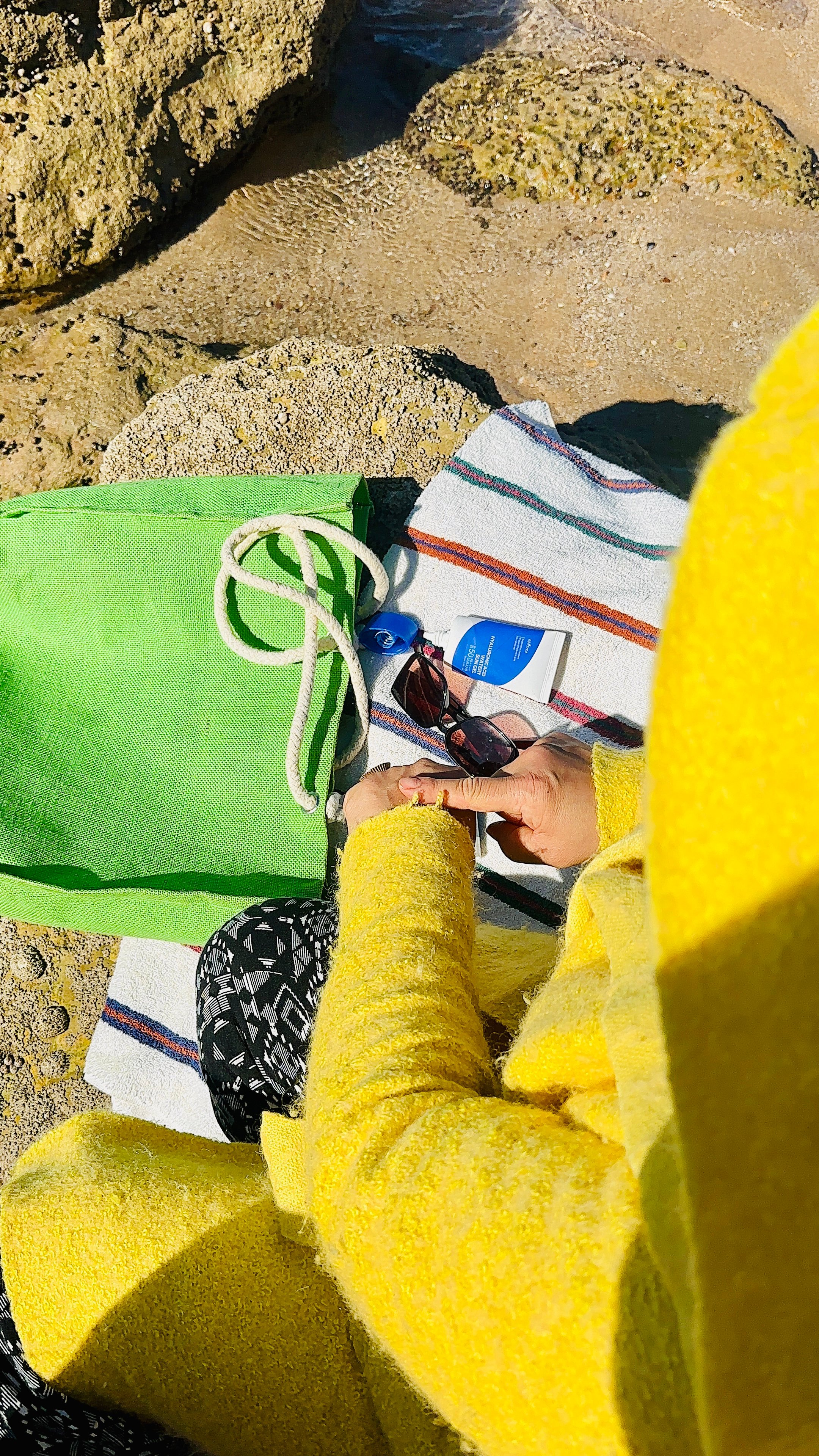 A person wearing a bright yellow sweater is sitting on a beach towel near the water, holding sunglasses with a green tote bag beside them.