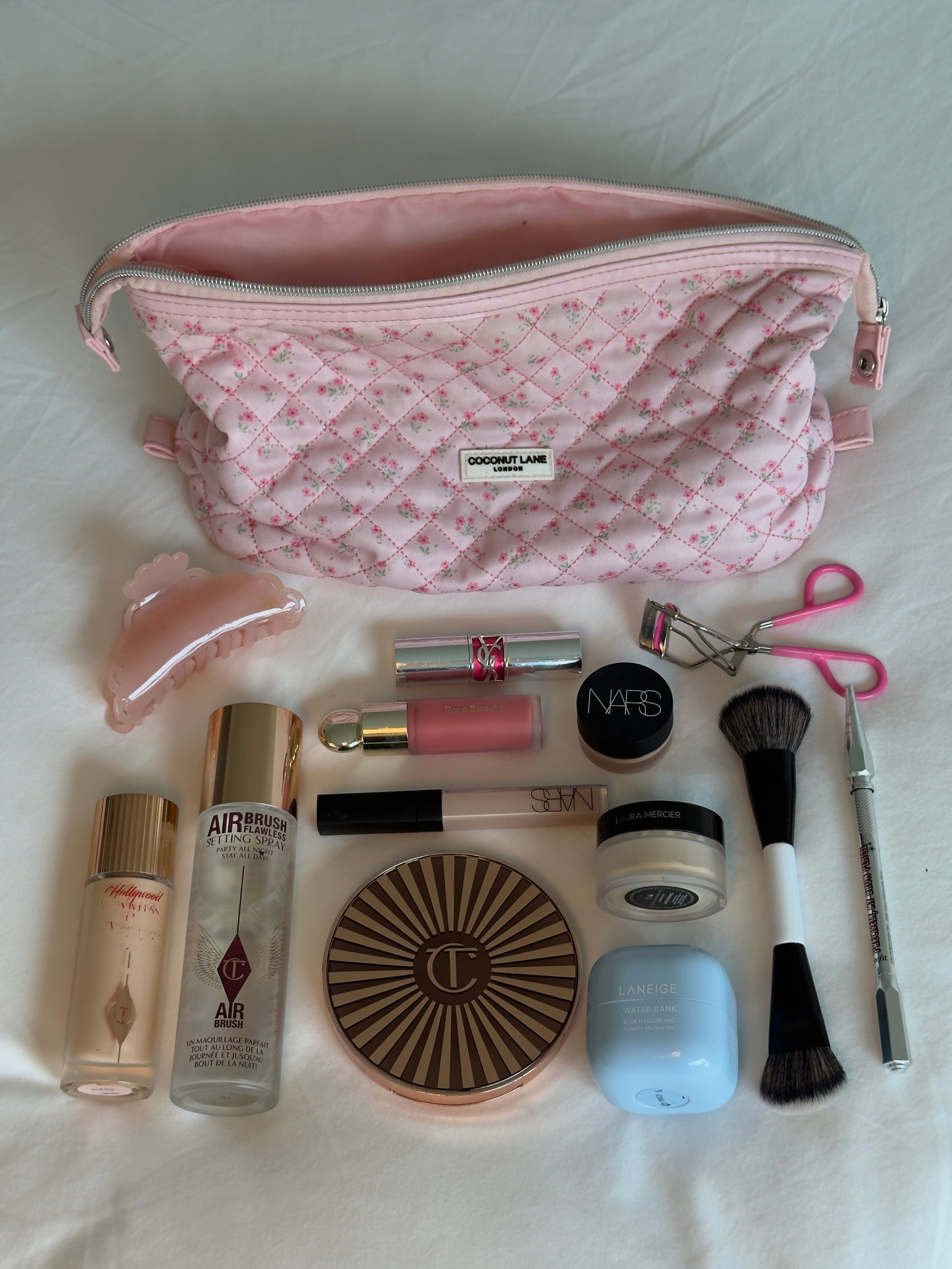 A neatly arranged collection of makeup products and tools displayed on a light background, featuring a pink quilted makeup bag.
