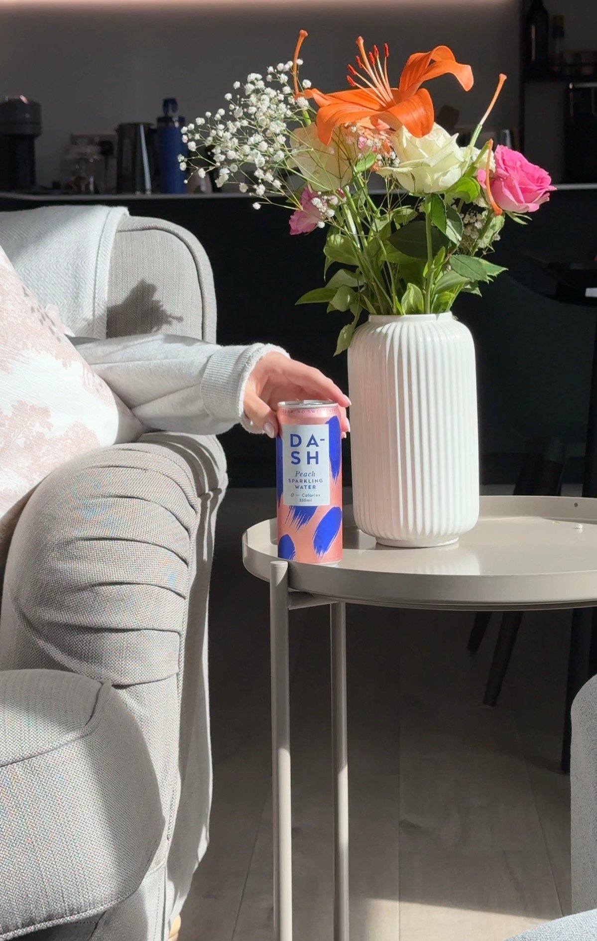 A hand holding a can of DASH beverage next to a vase of colorful flowers on a small table in a cozy setting.