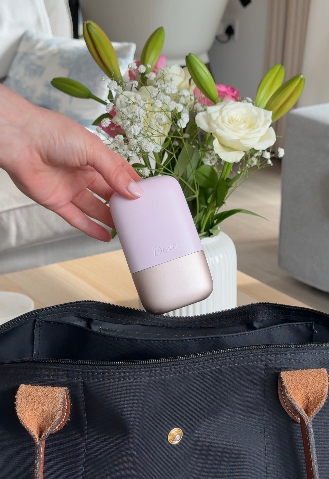 A hand holding a pink and beige portable device above an open black bag, with a bouquet of flowers in the background.