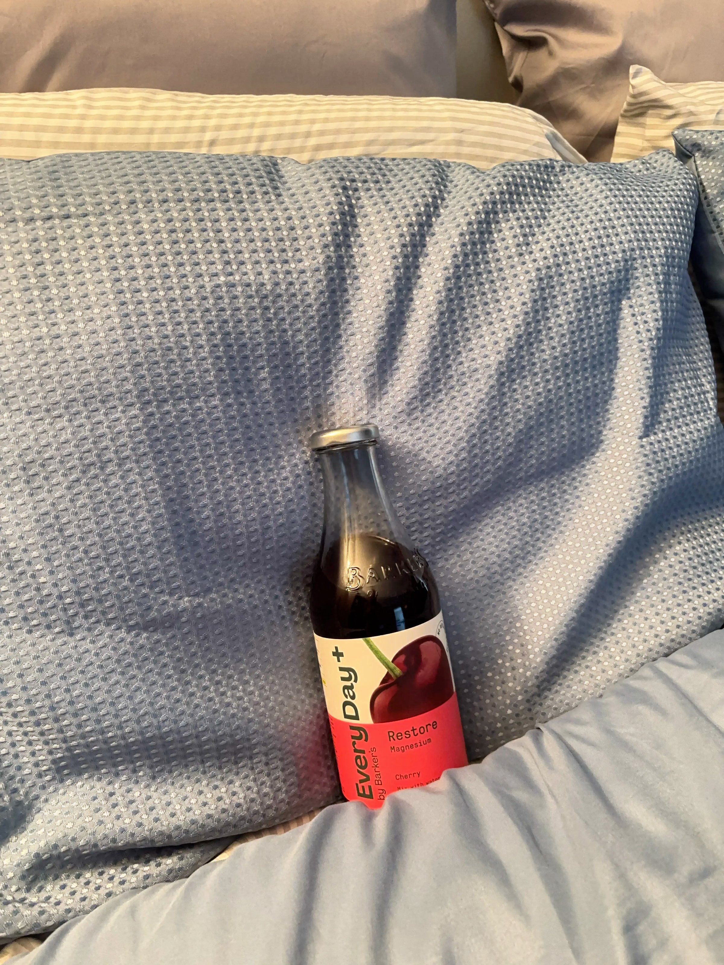 A bottle of cherry soda resting on a bed with blue pillows and bedding.
