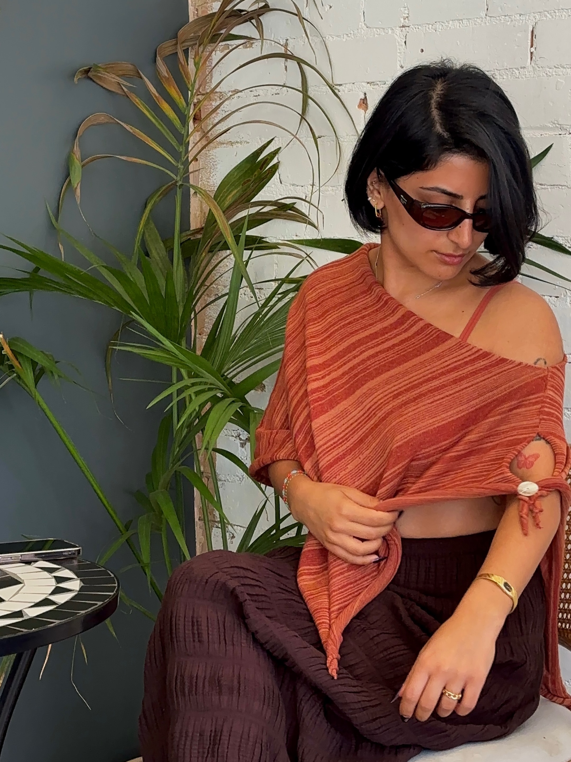 A woman wearing a stylish orange wrap top and sunglasses, seated beside a potted plant and a patterned table.