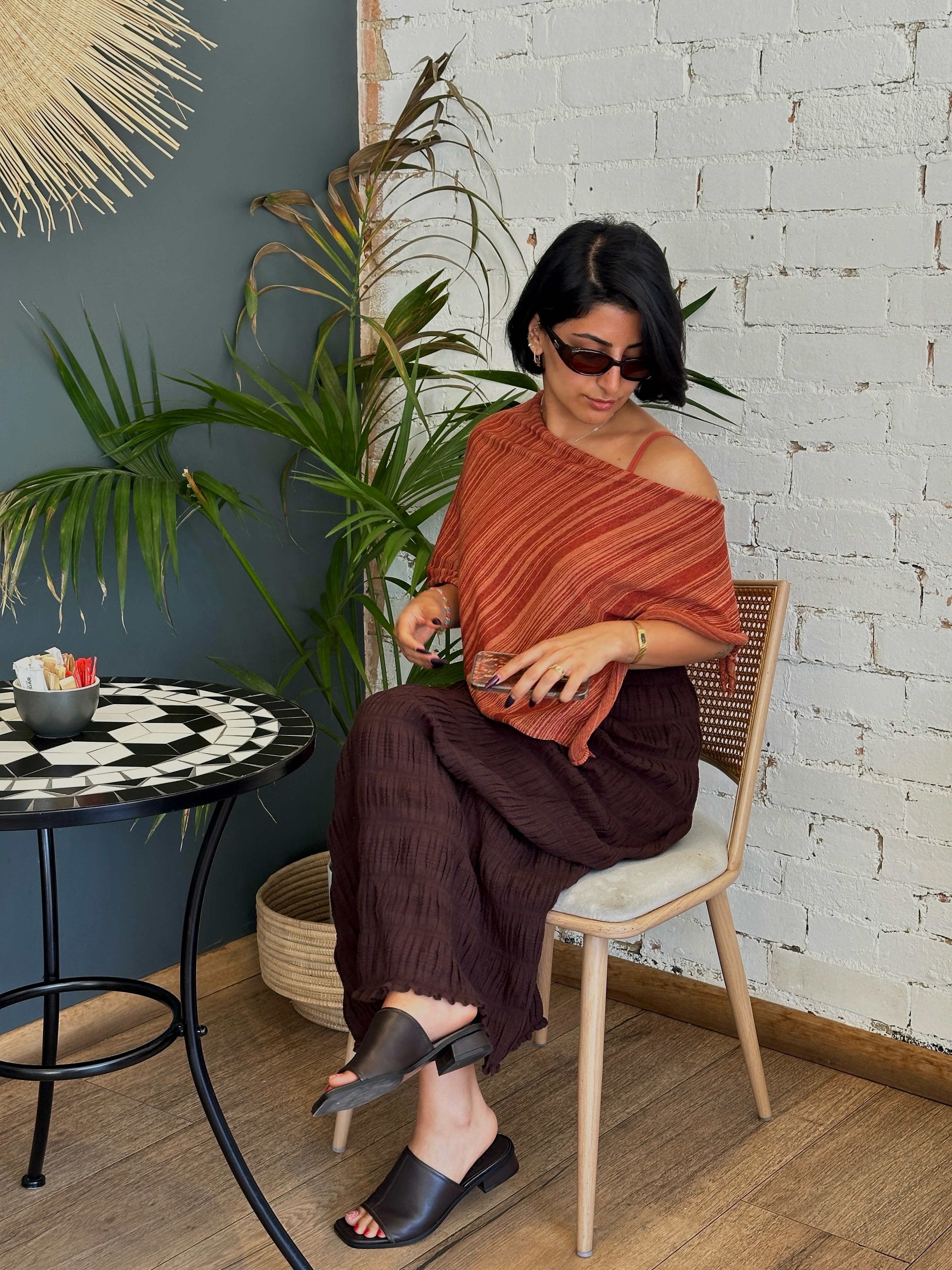 A woman sits at a small table with a checkered surface, wearing a stylish off-shoulder top and sunglasses, surrounded by greenery and a textured wall.
