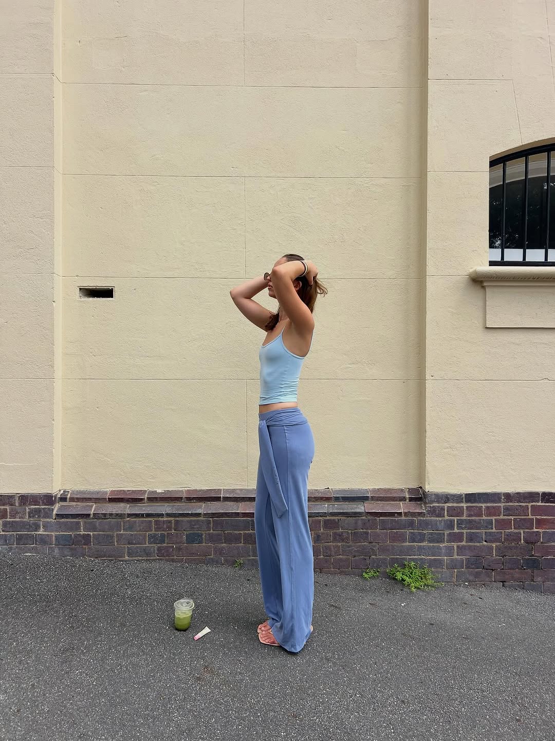 A woman stands sideways against a light-colored wall, adjusting her hair. She wears a light blue tank top and loose-fitting blue pants, with a green drink on the ground beside her.